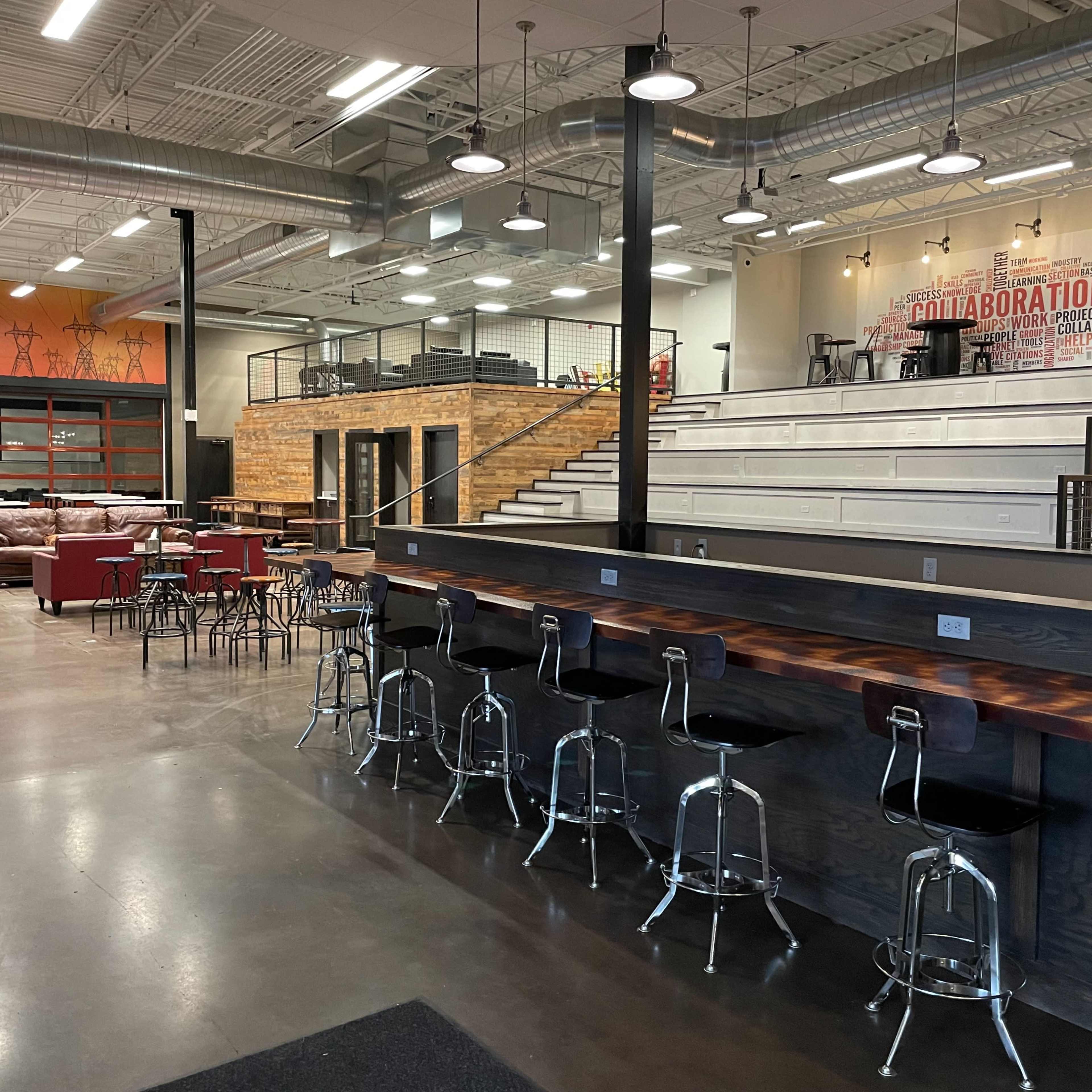 An industrial-style interior featuring a spacious open area with a bar counter, seating arrangements, and a tiered seating section at the rear.