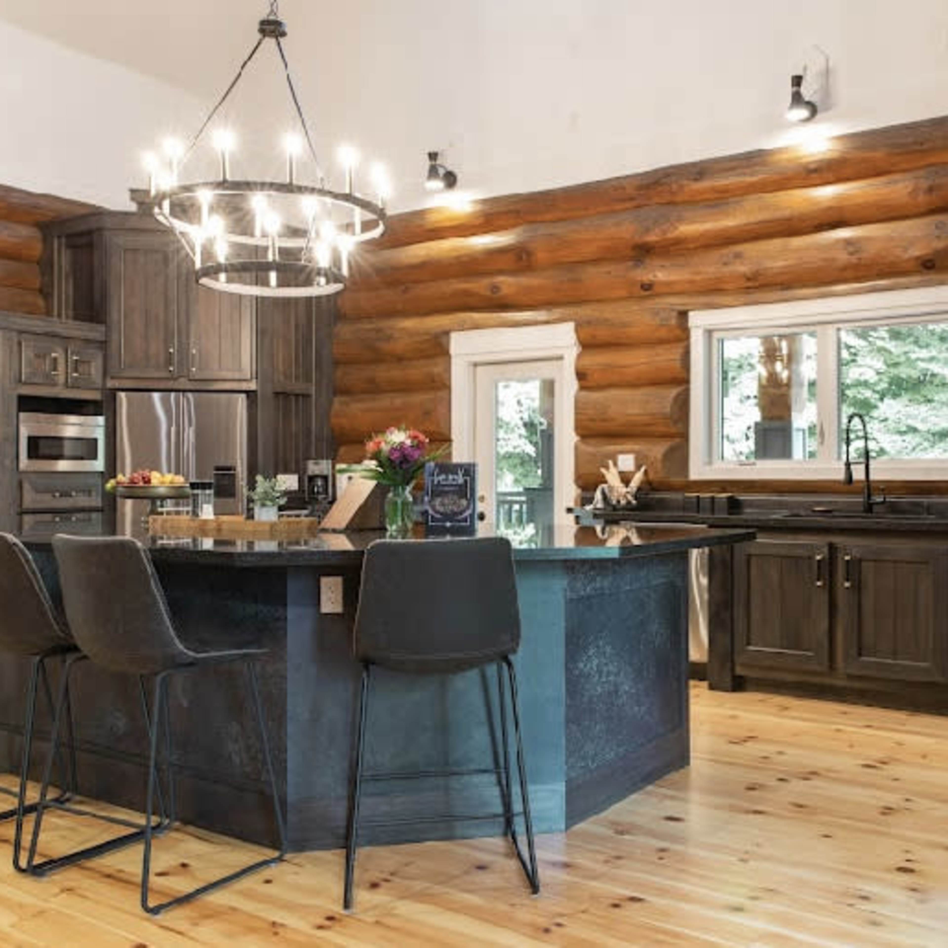 A modern kitchen with dark wood cabinetry, a large island with seating, and a spacious area featuring a chandelier and large windows.