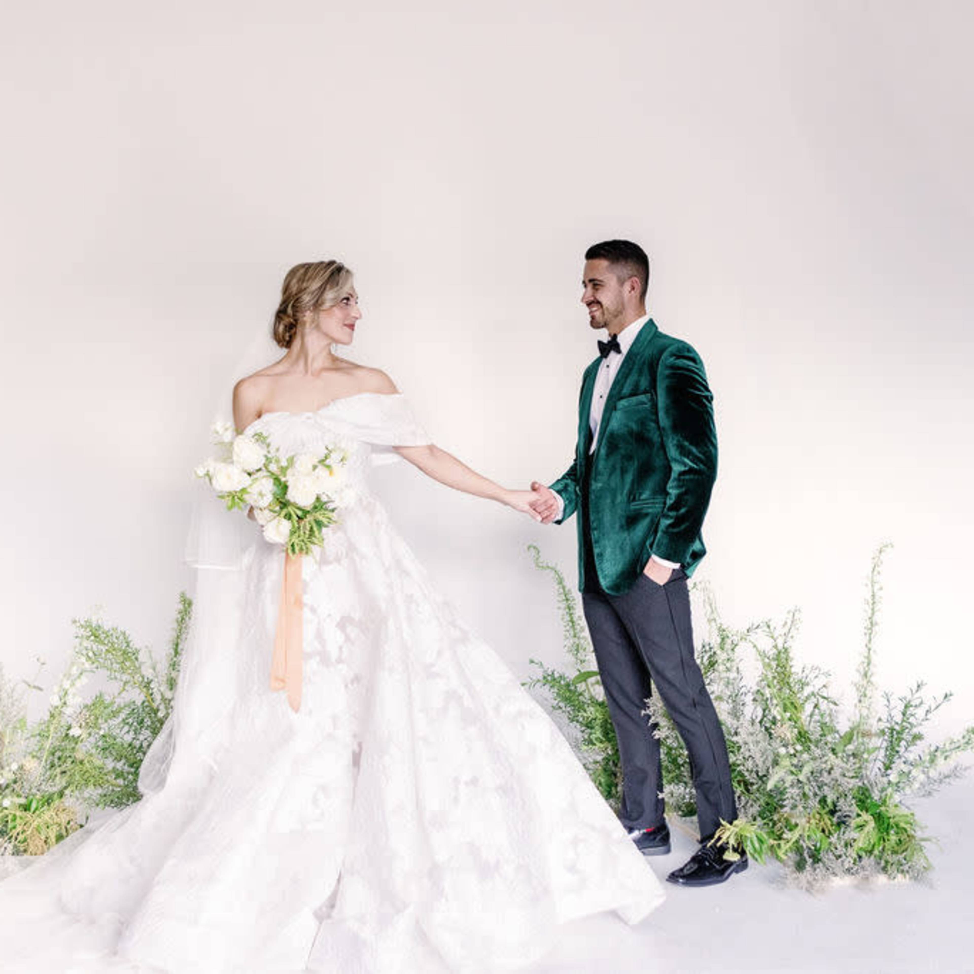 A bride in a white off-the-shoulder gown holds hands with a groom in a green velvet jacket against a simple backdrop adorned with greenery.