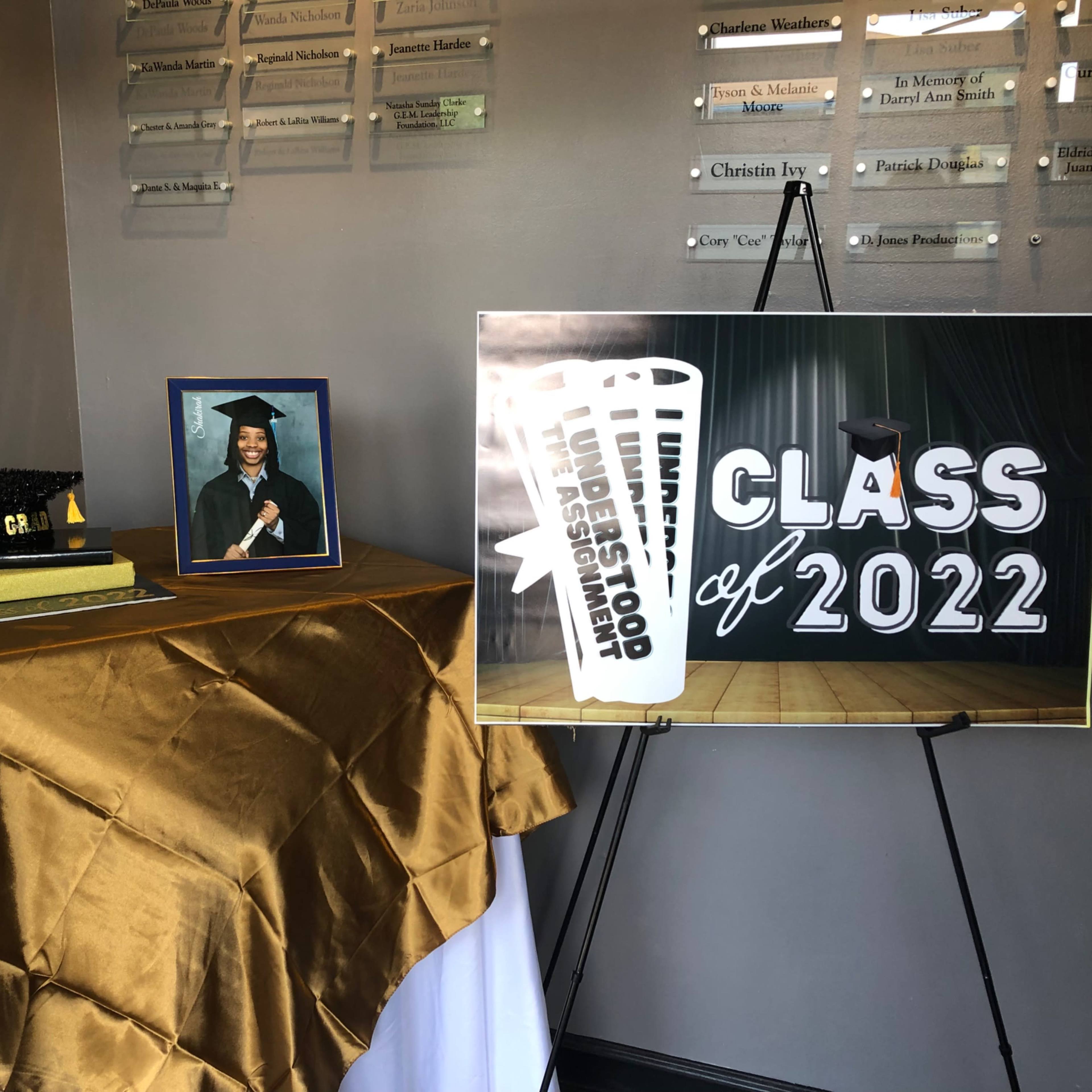 A graduation display featuring a framed portrait of a graduate, a graduation cap, a decorative book, and a large sign that reads "Class of 2022" on a table covered with a golden cloth.