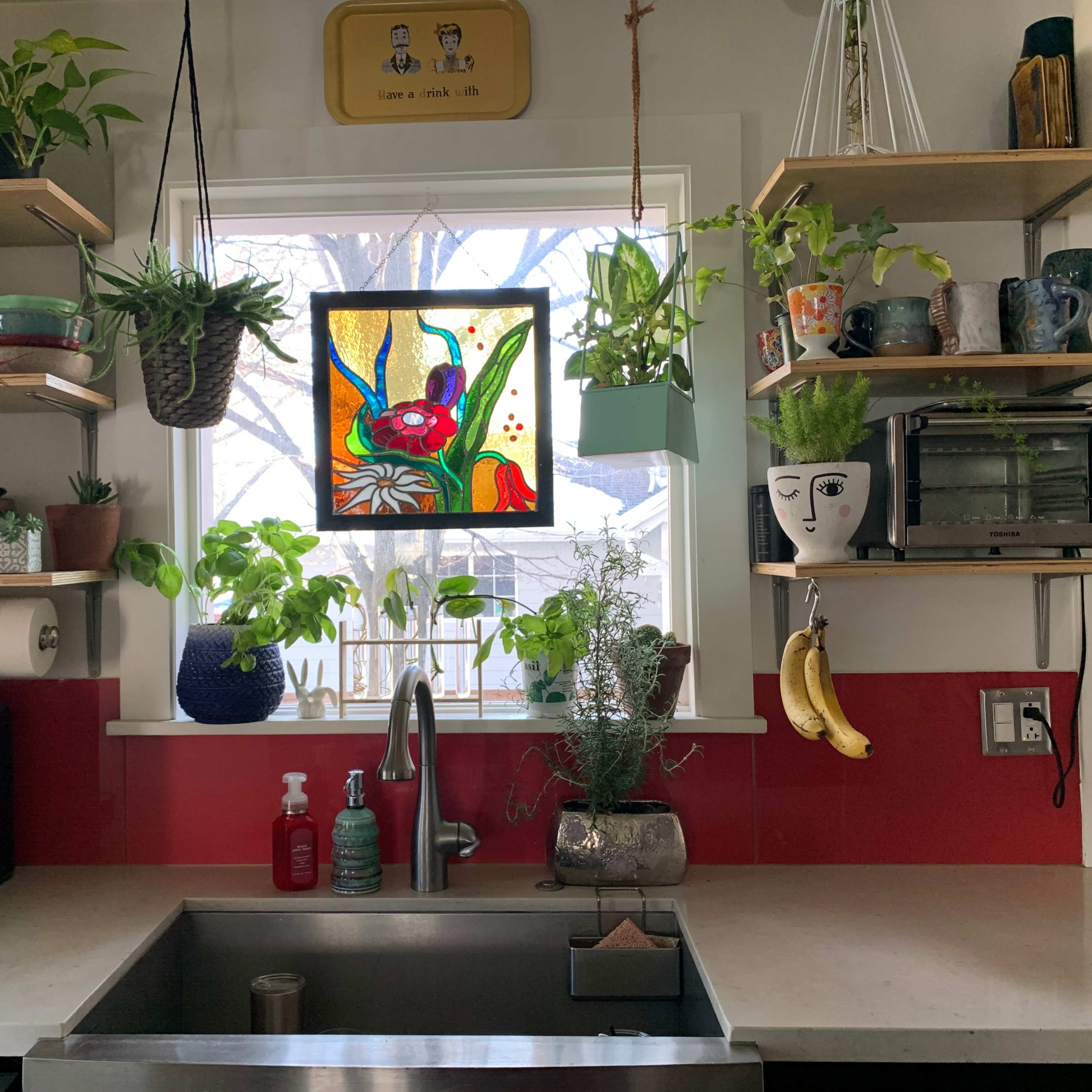 A kitchen sink area with shelves holding plants, various mugs, and an art piece, framed by a window overlooking a garden.
