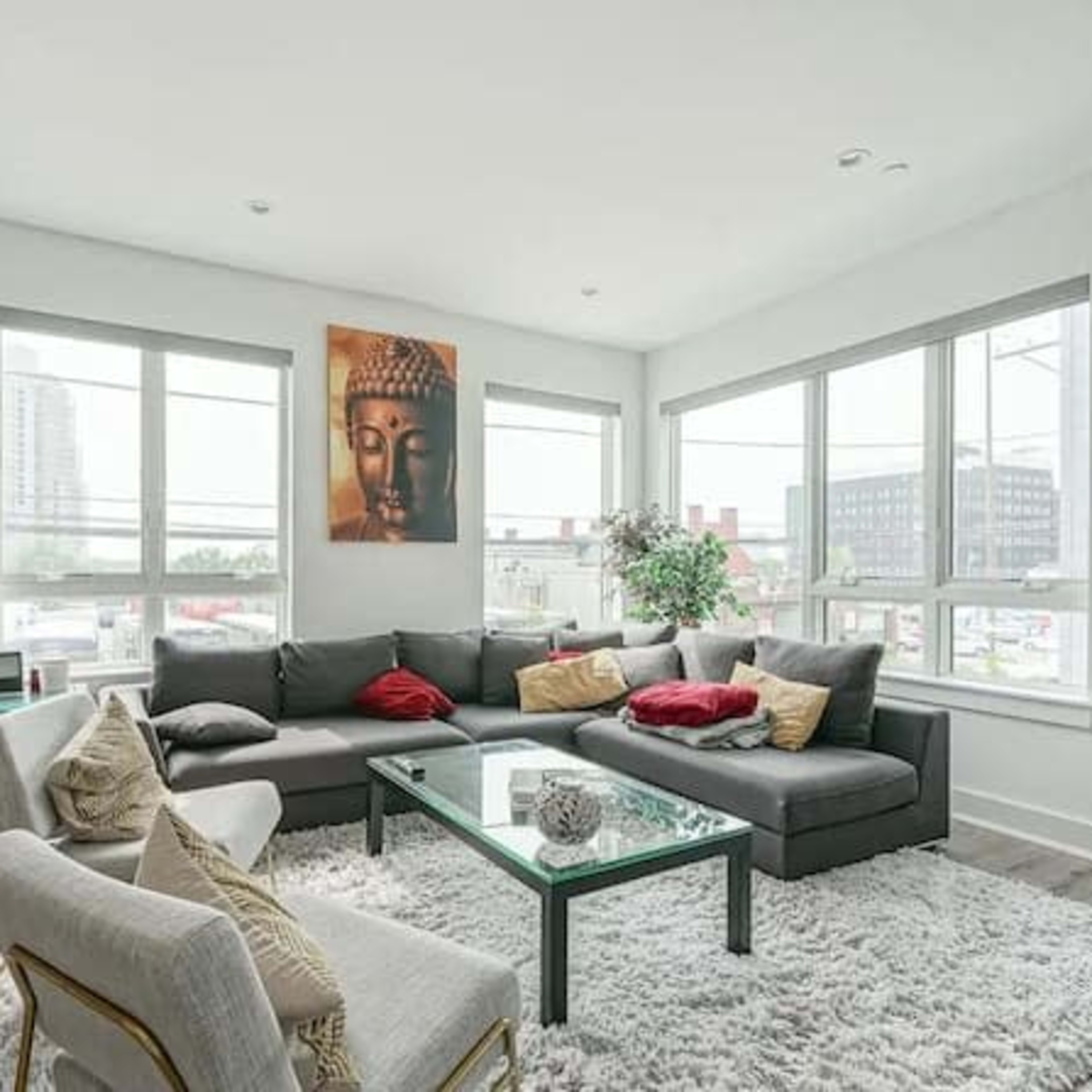 The living room features a sectional sofa with decorative pillows, a glass coffee table on a plush rug, and large windows that provide natural light.