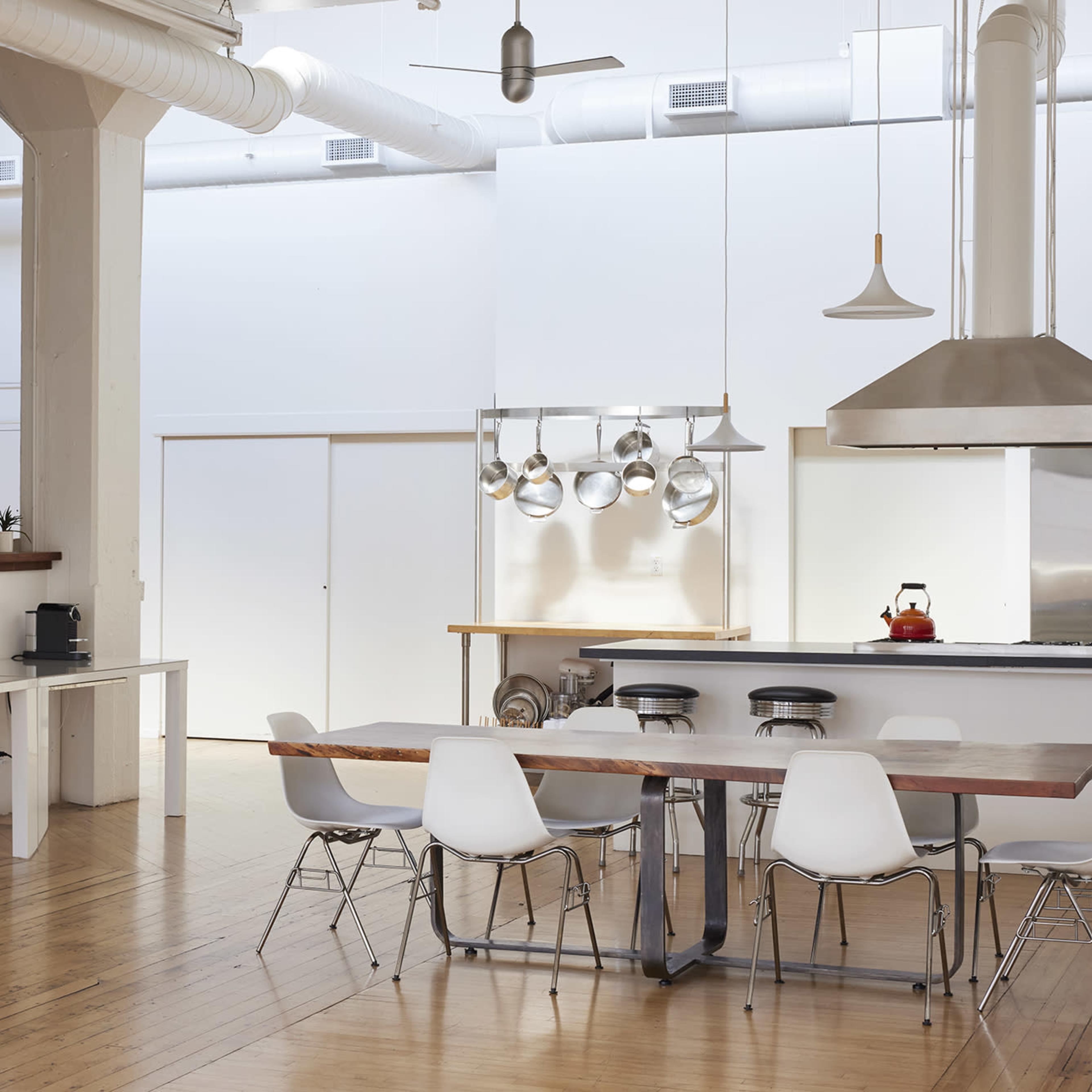 A bright and open kitchen and dining area features a large wooden table surrounded by white chairs, with stainless steel appliances and hanging pots visible in the background.