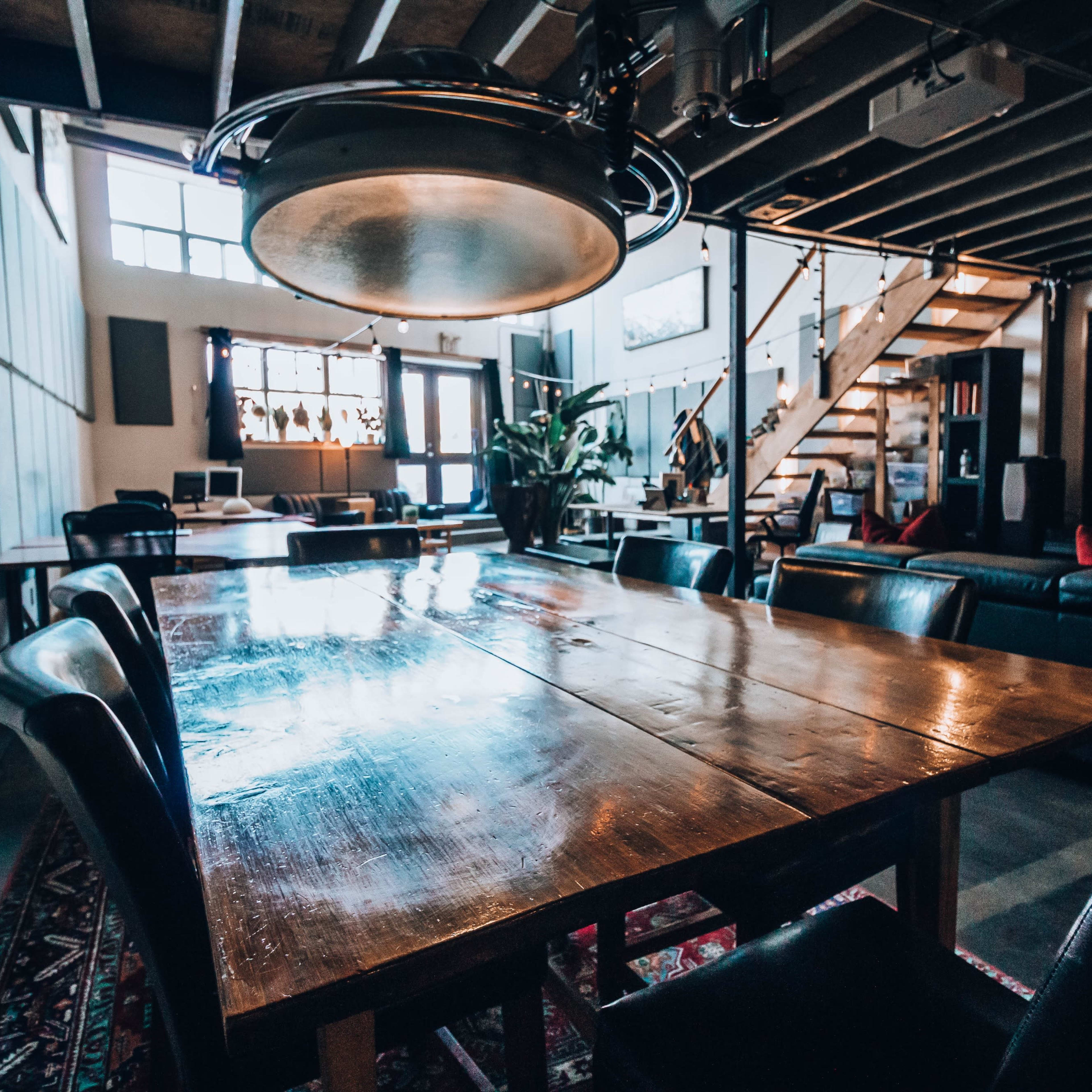 A spacious, modern living area features a large wooden dining table surrounded by black leather chairs, with a staircase and a multimedia setup in the background.