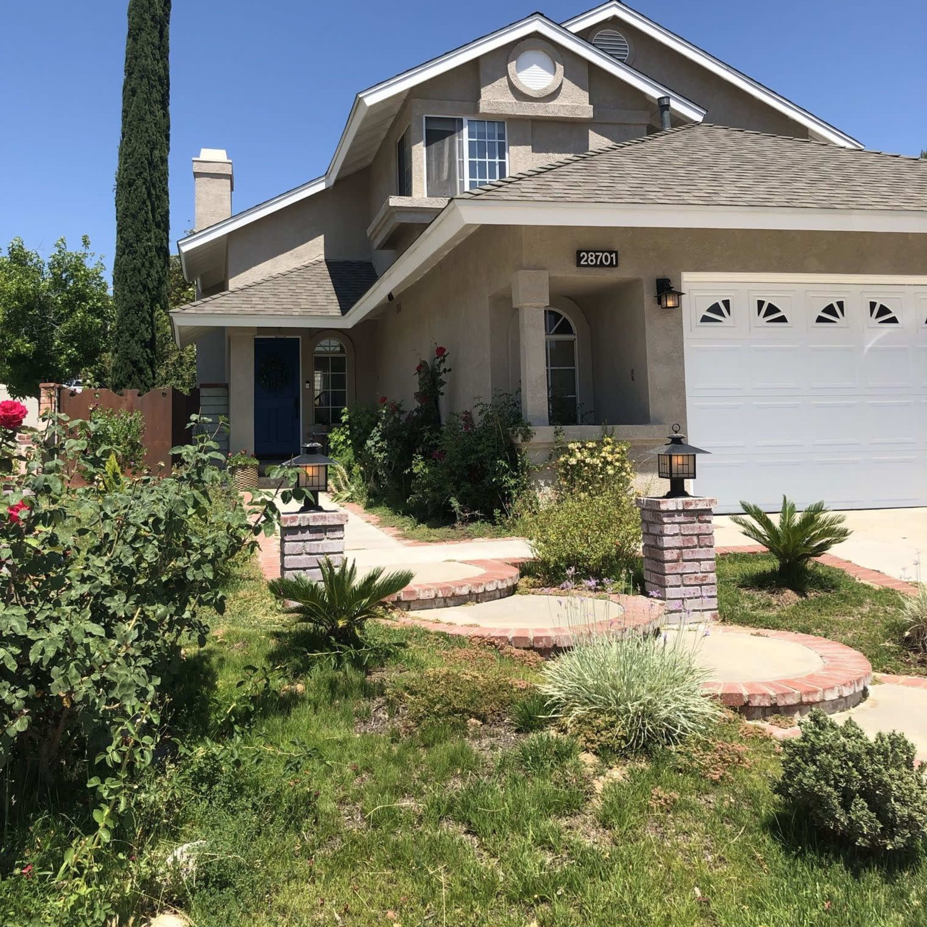 A two-story house with a gray exterior, a garage, and landscaped front yard featuring pathways, shrubs, and a tall cypress tree.