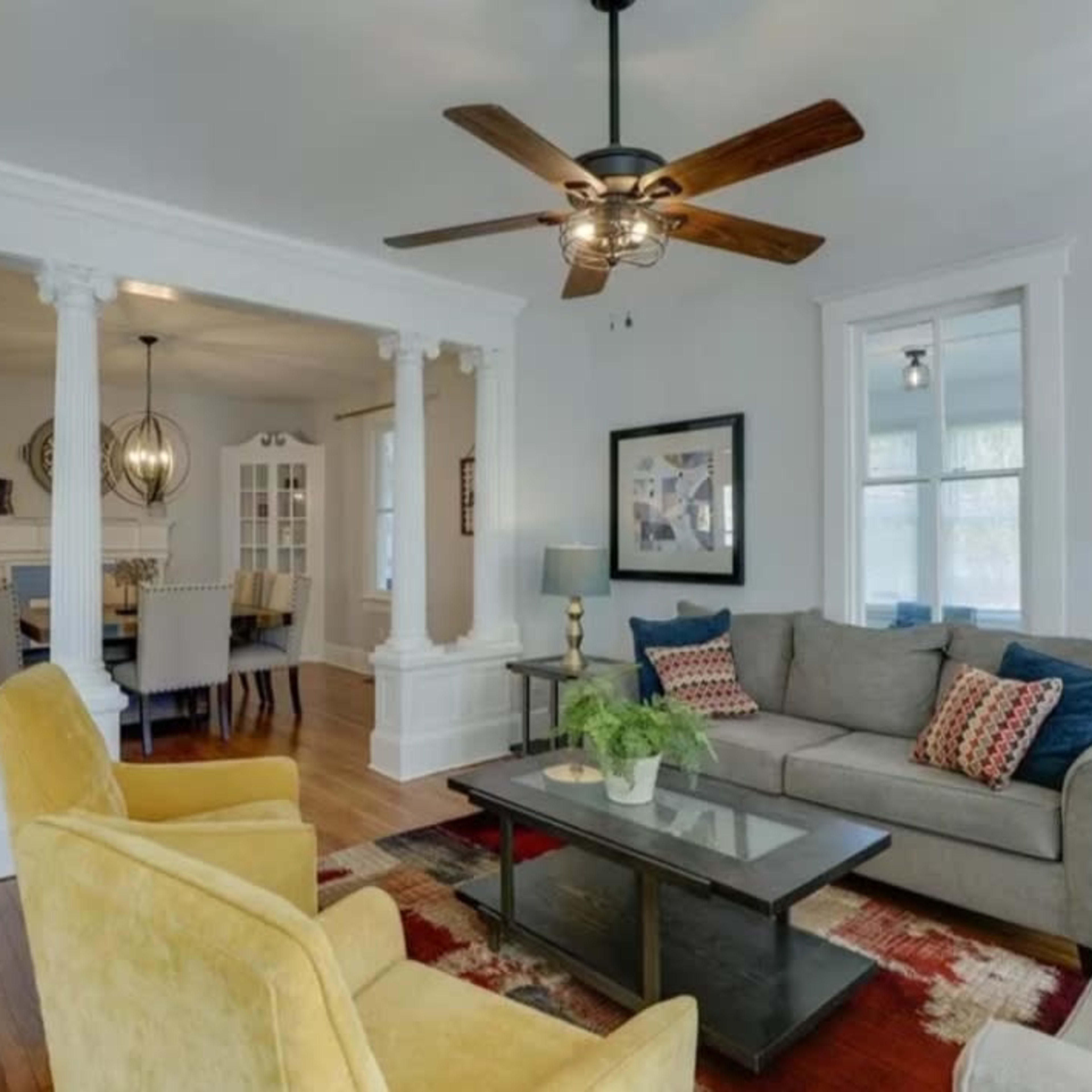 A living room with a sofa and yellow chairs, featuring a ceiling fan and a view into a dining area through a set of white columns.