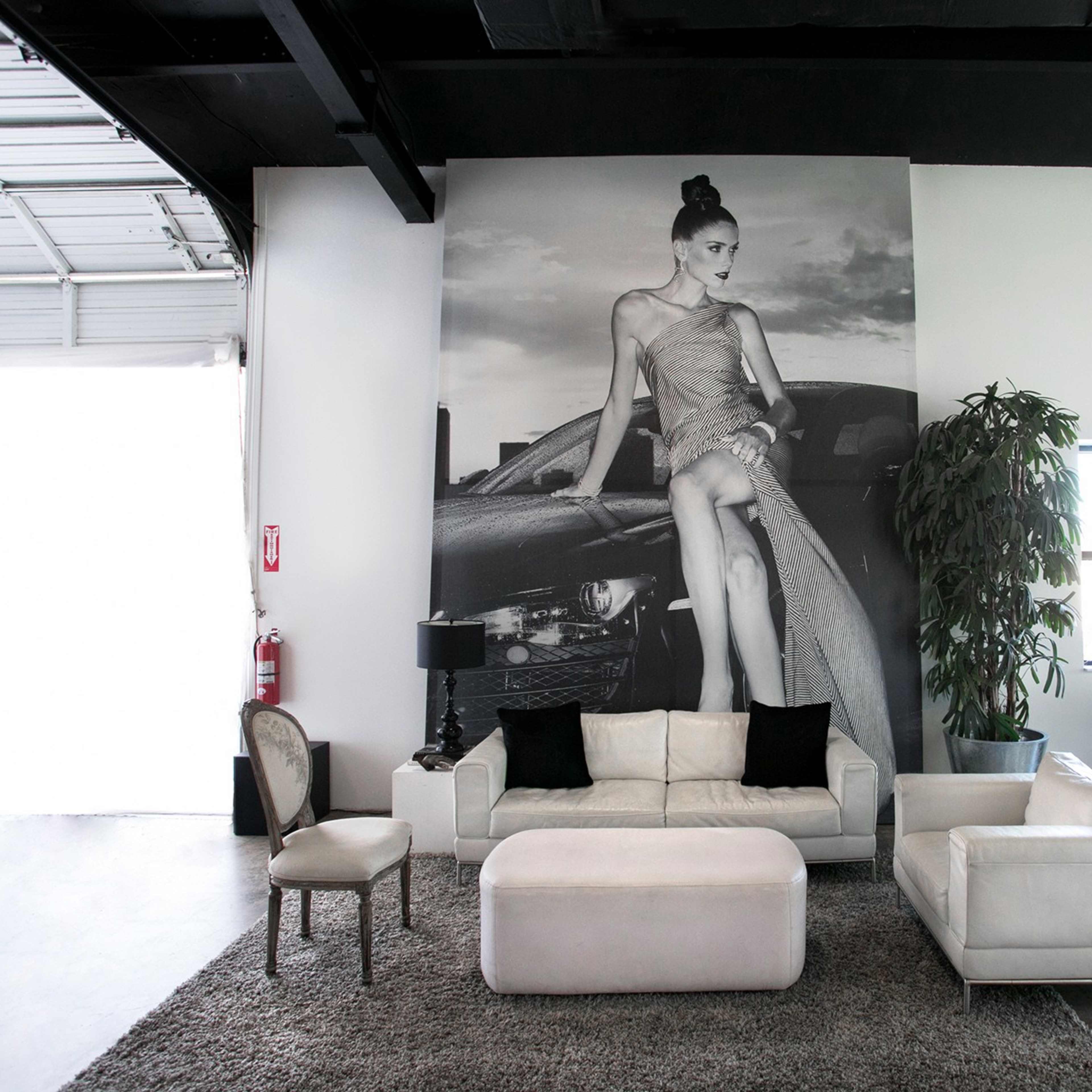 The scene features a modern lounge area with a white sofa, an ottoman, and a large black-and-white mural of a woman next to a car on the wall.