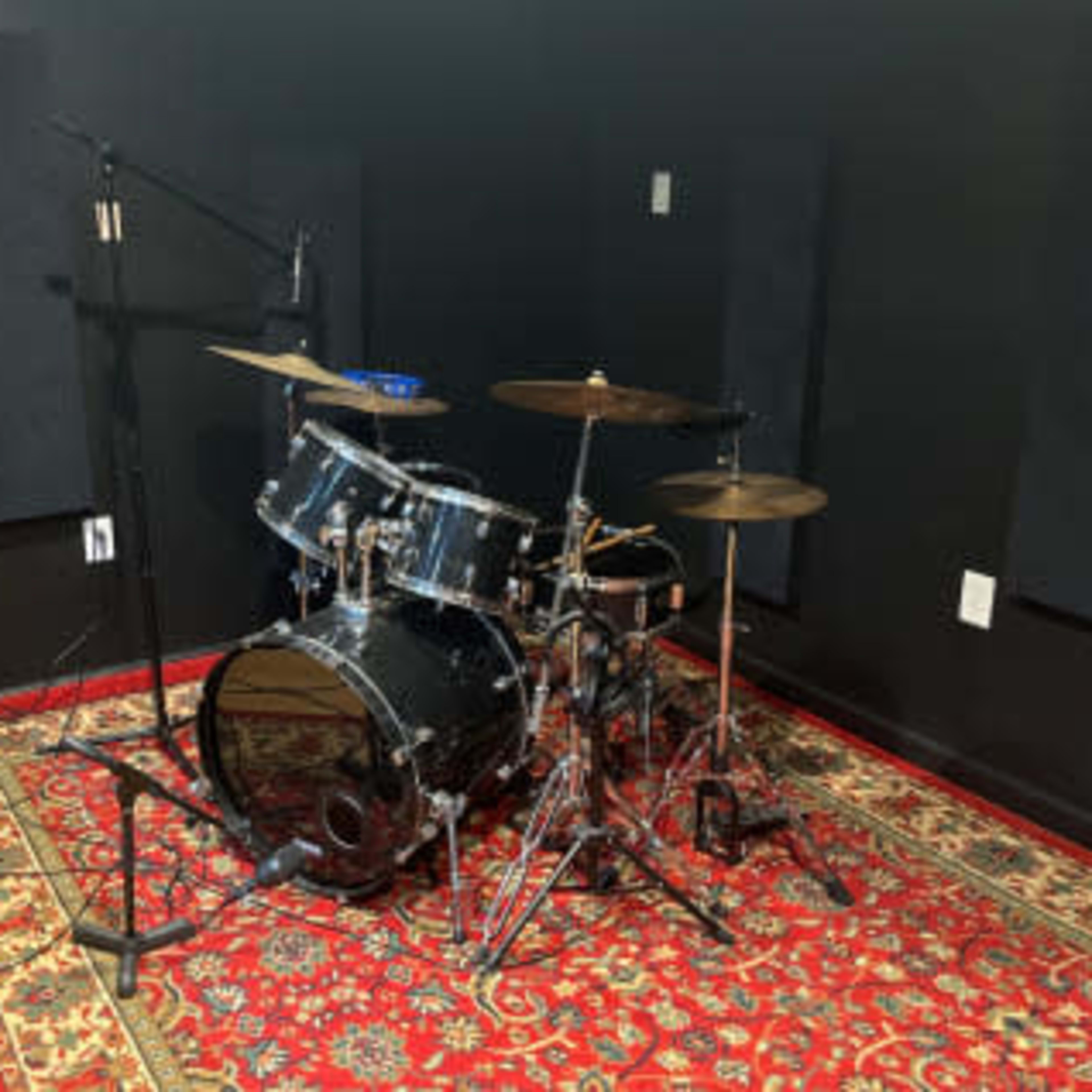 A black-painted room features a drum set positioned on a patterned rug, with sound-absorbing panels on the walls.