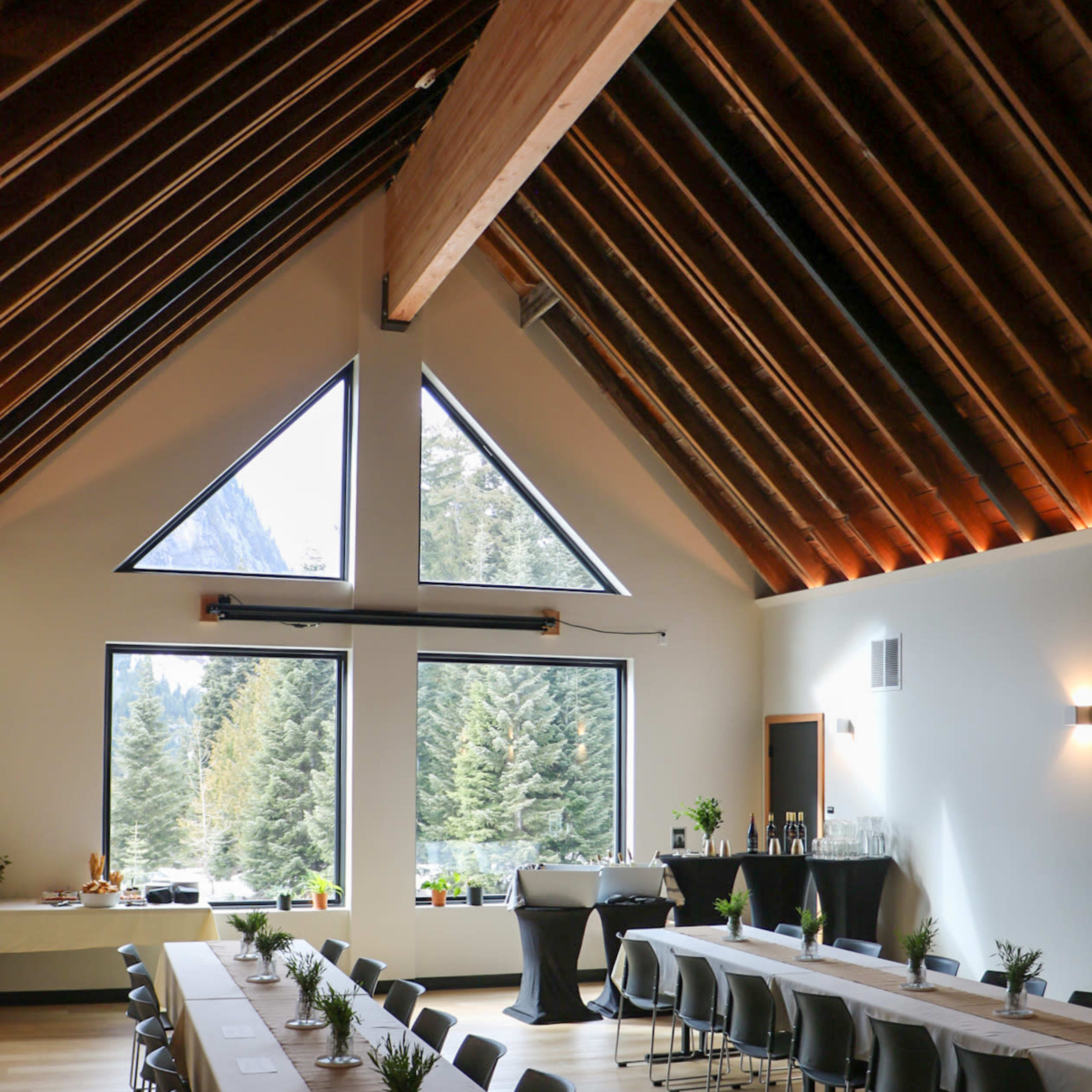 A spacious indoor event space with large windows showcasing a view of trees, featuring two long tables set up for a gathering.