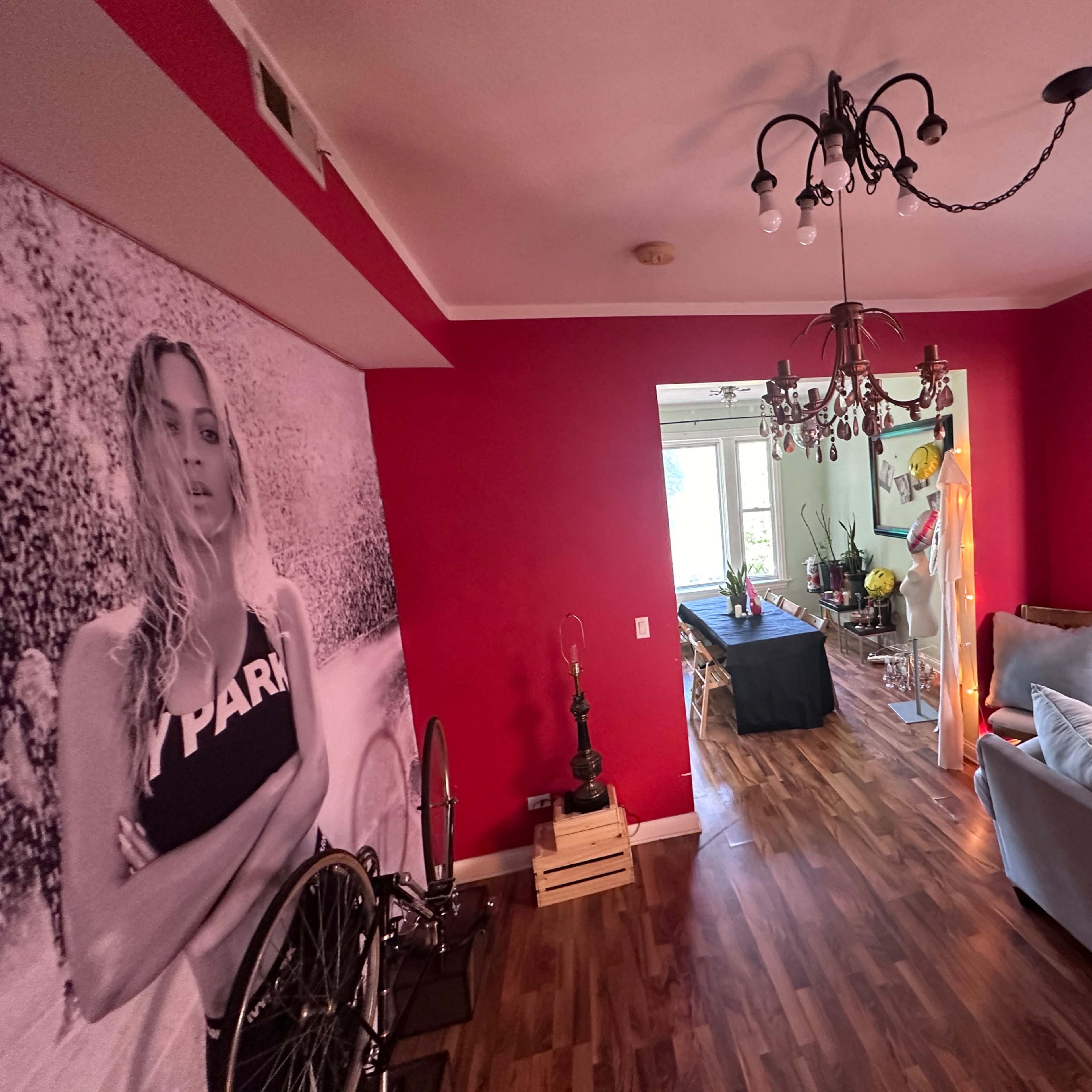 A living area with red walls, a large black and white photograph of a woman on one wall, and a view through an archway leading to another room with a table and decorations.