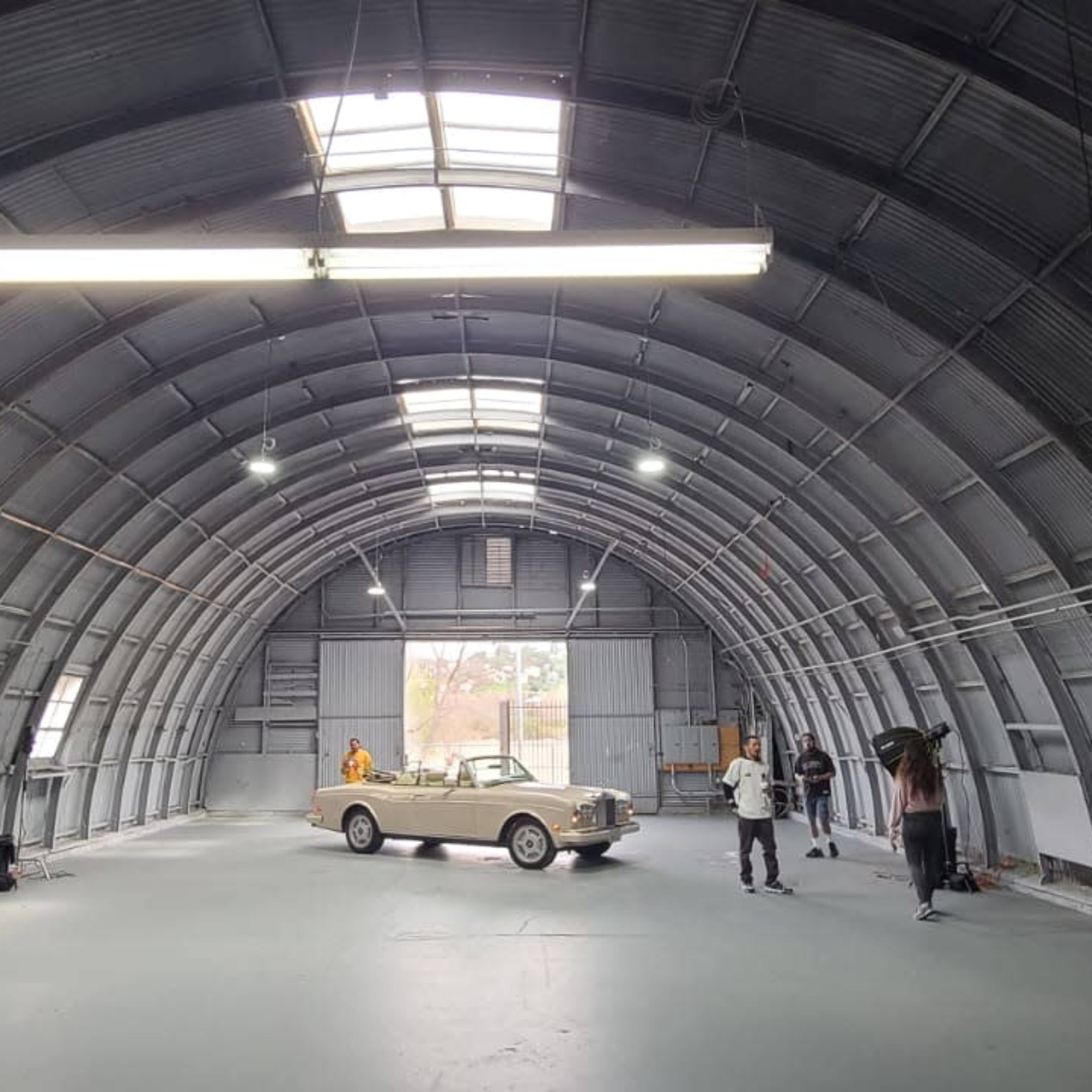 A large, arched metal building houses a few people and a car on a gray floor.