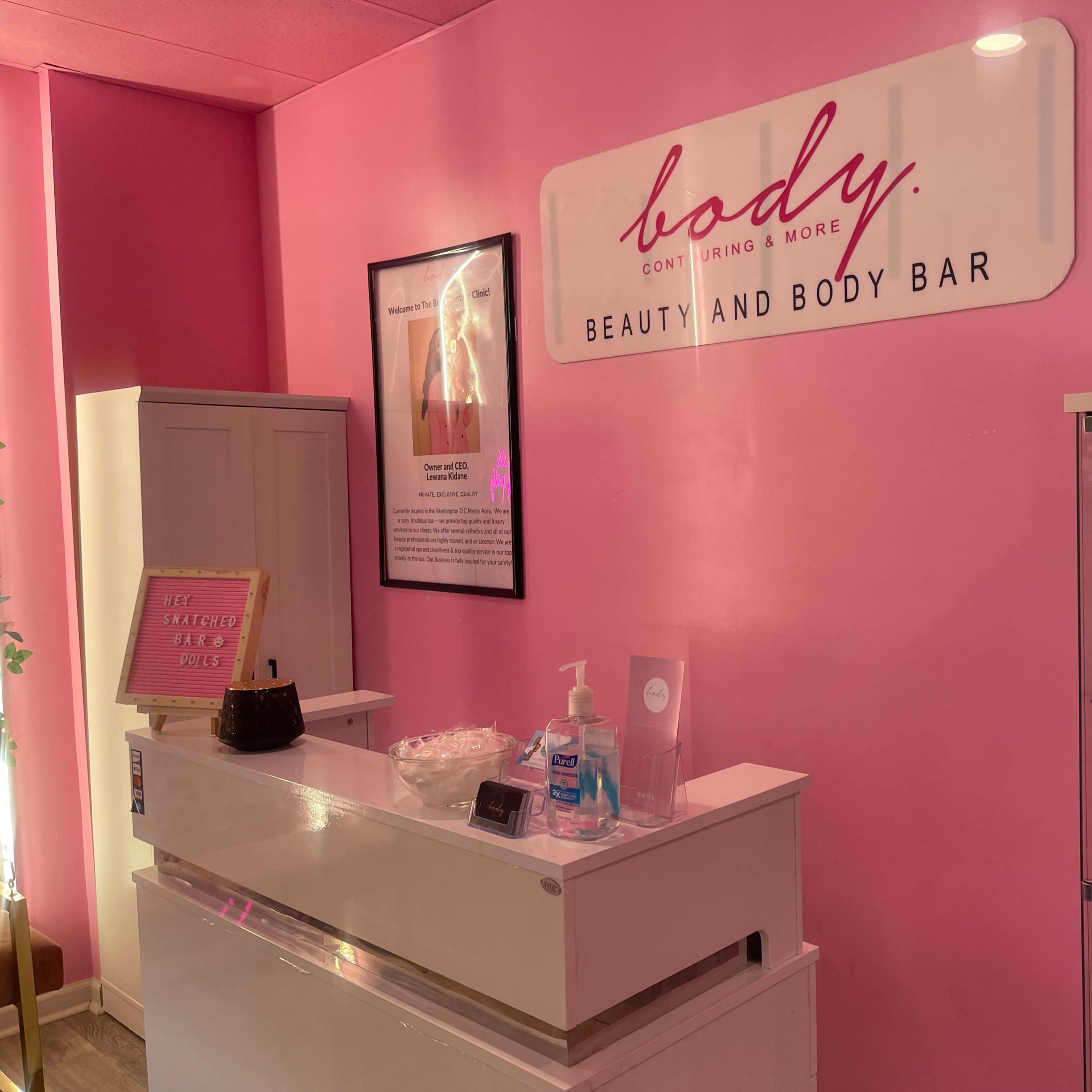 The interior of a beauty and body bar with a pink wall, a reception desk, and display shelves featuring beauty products.