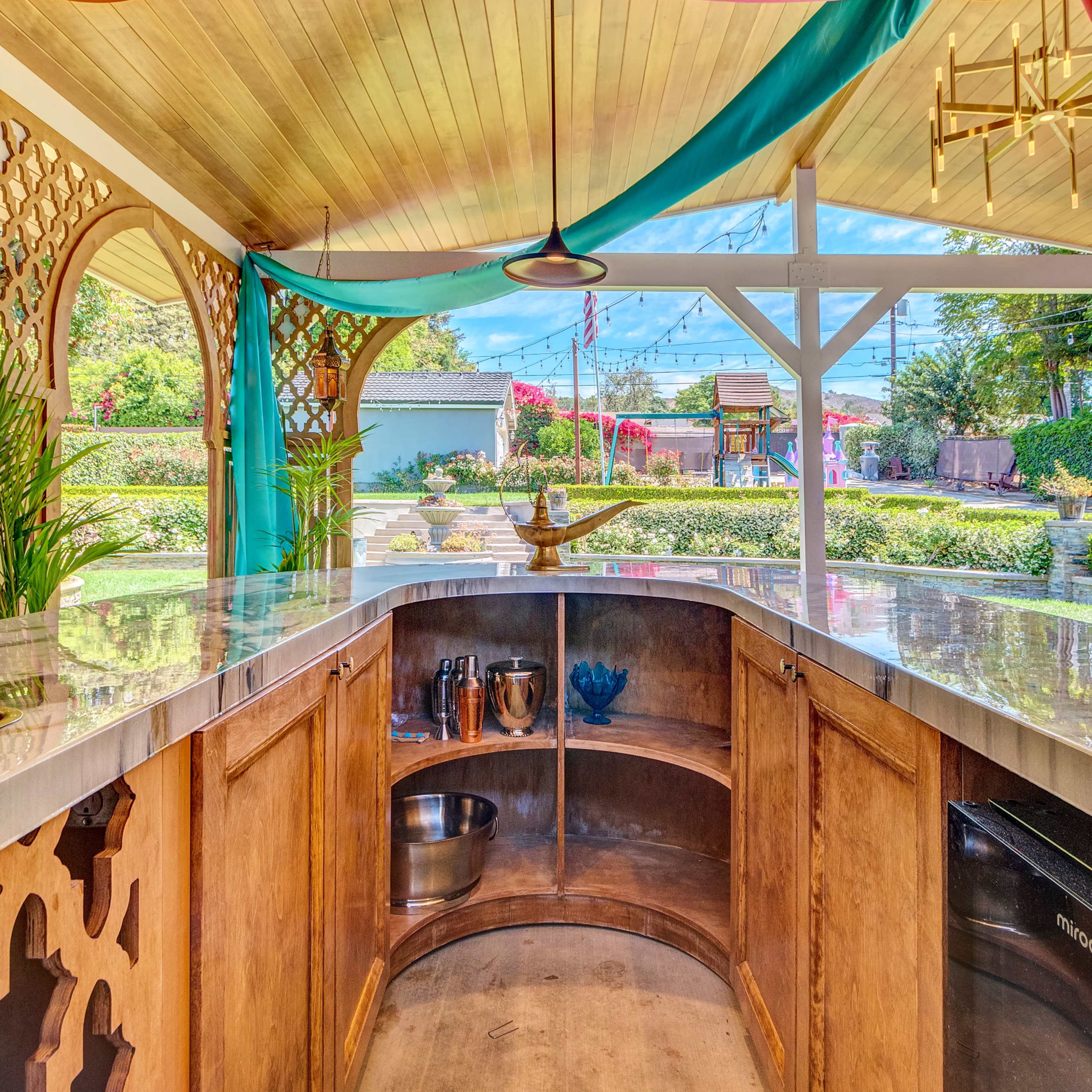 A bar area with a curved wooden counter, decorative lattice panels, and colorful fabric draping from the ceiling, set in a garden-like environment.