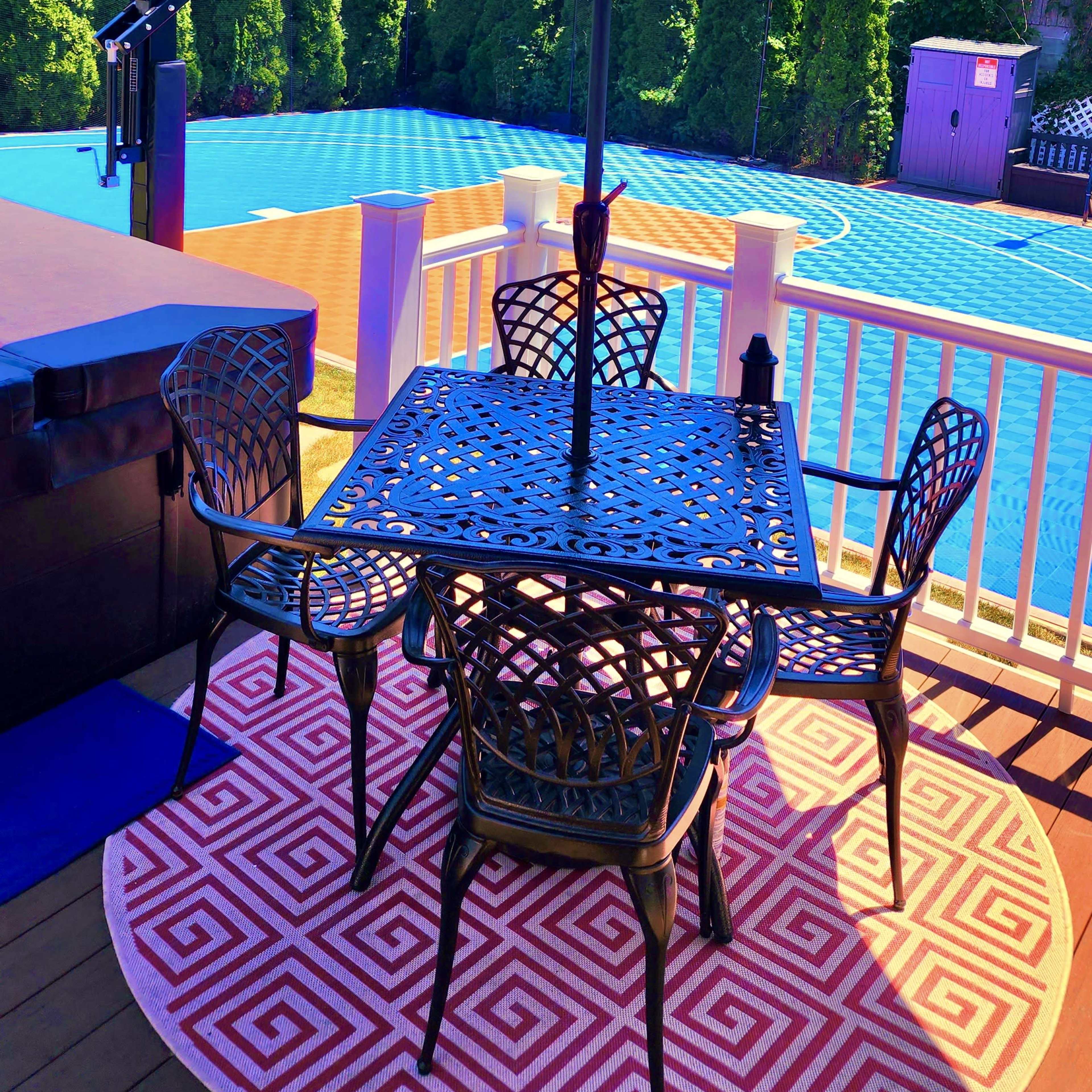 A blue umbrella shades a black outdoor table with four chairs on a patio overlooking a swimming pool surrounded by greenery.