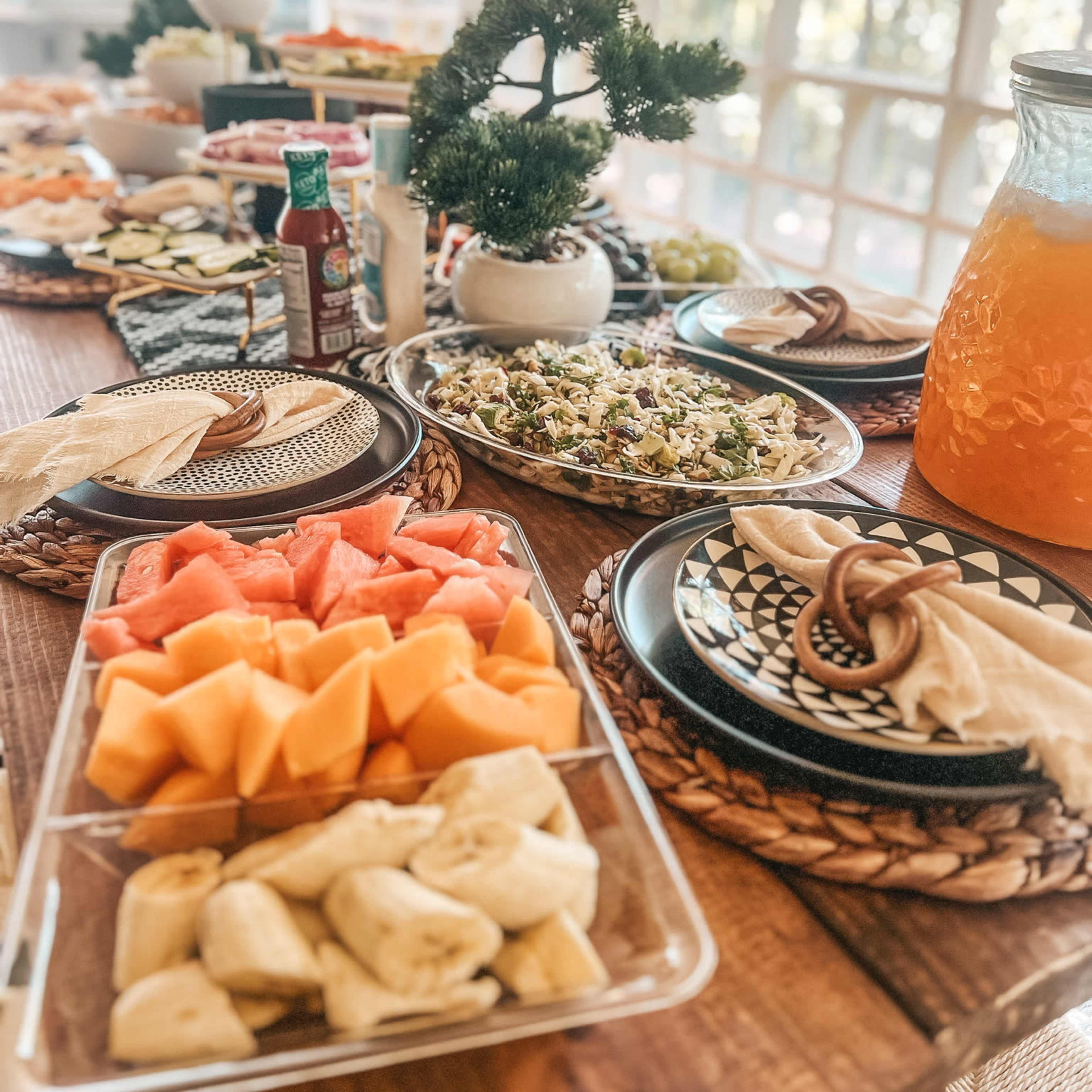 A table is filled with a variety of dishes, including fruits, salads, and beverages, arranged for a gathering in a bright, sunlit room.