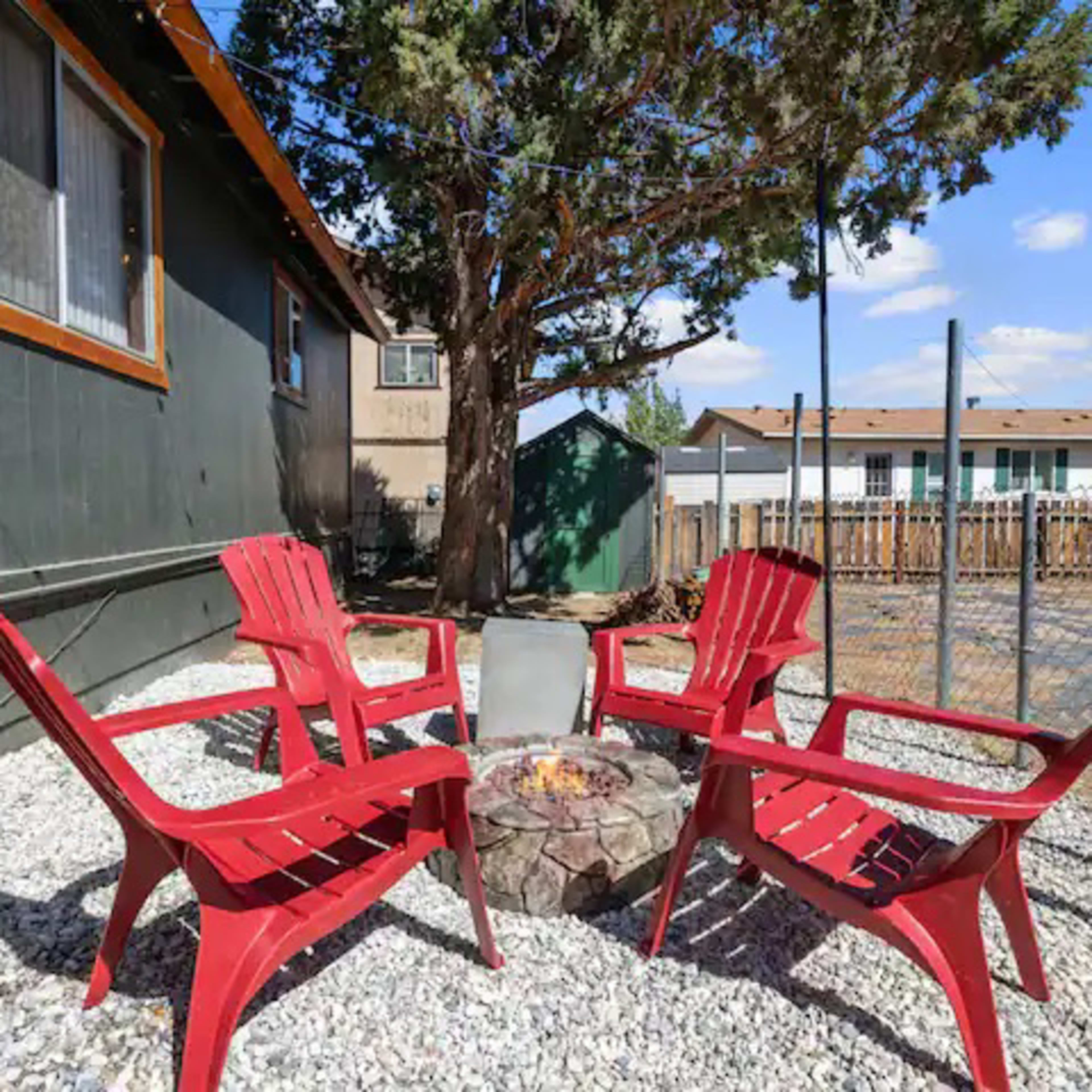 A circular fire pit surrounded by four red Adirondack chairs sits in a gravel area next to a house and a fenced yard.