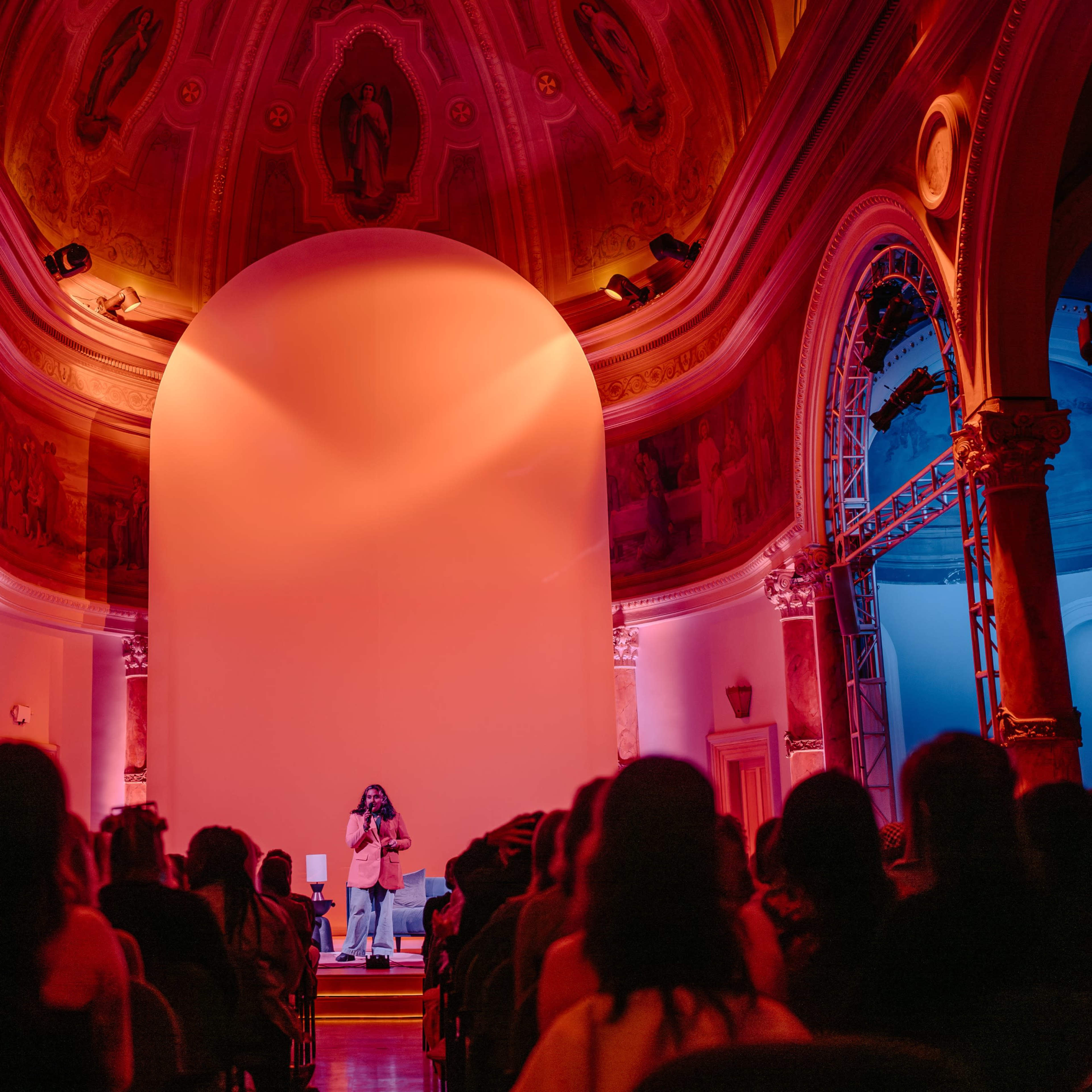 A speaker addresses an audience in a vividly lit hall with ornate architecture.
