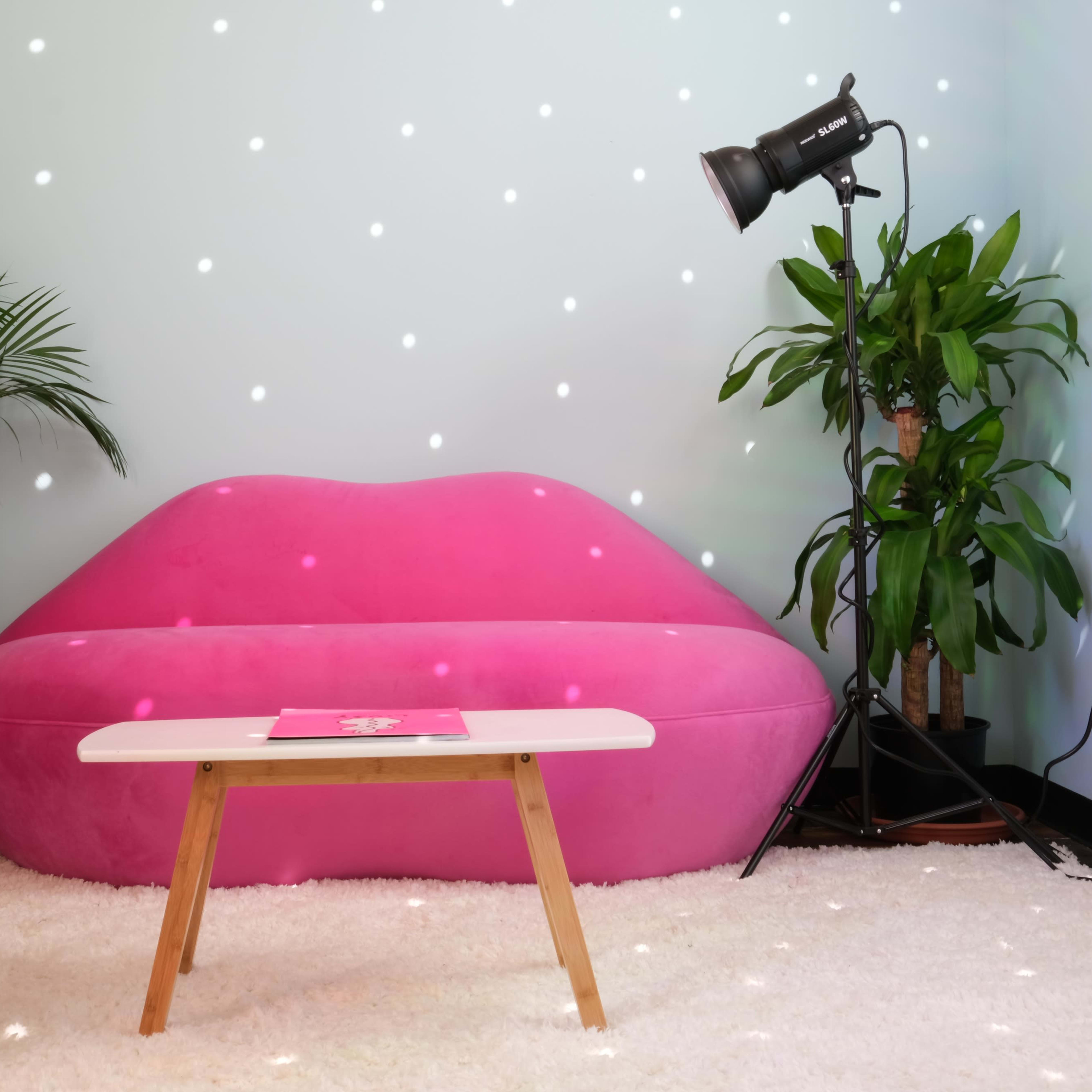 A pink lips-shaped sofa positioned against a light blue wall, accompanied by two plants, a small white table, and a spotlight.