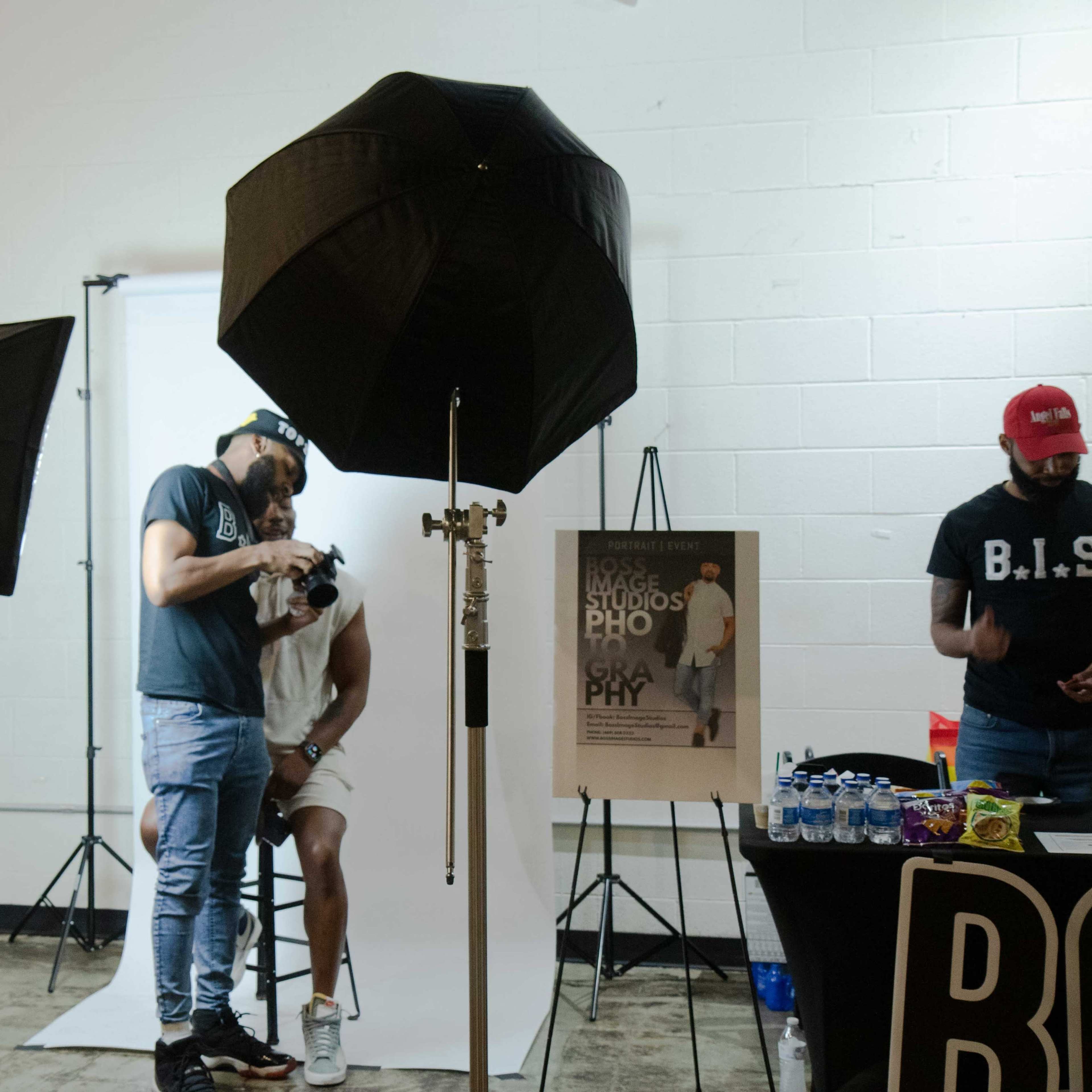 A photographer adjusts a camera while sitting beside a model in front of two softbox lights and a backdrop.