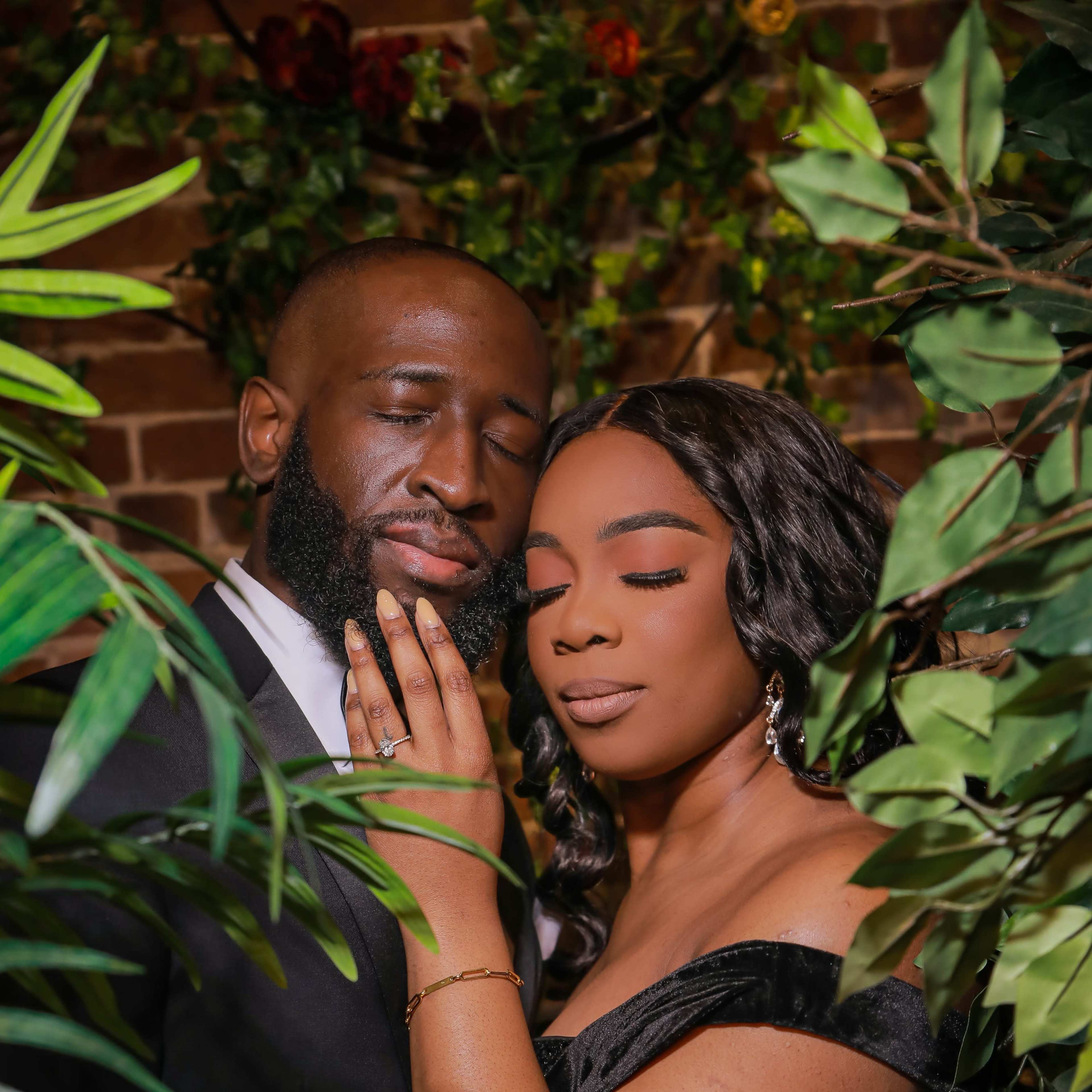 A couple poses together amidst lush greenery and flowers, with the man gently touching the woman's face as they share an intimate moment.
