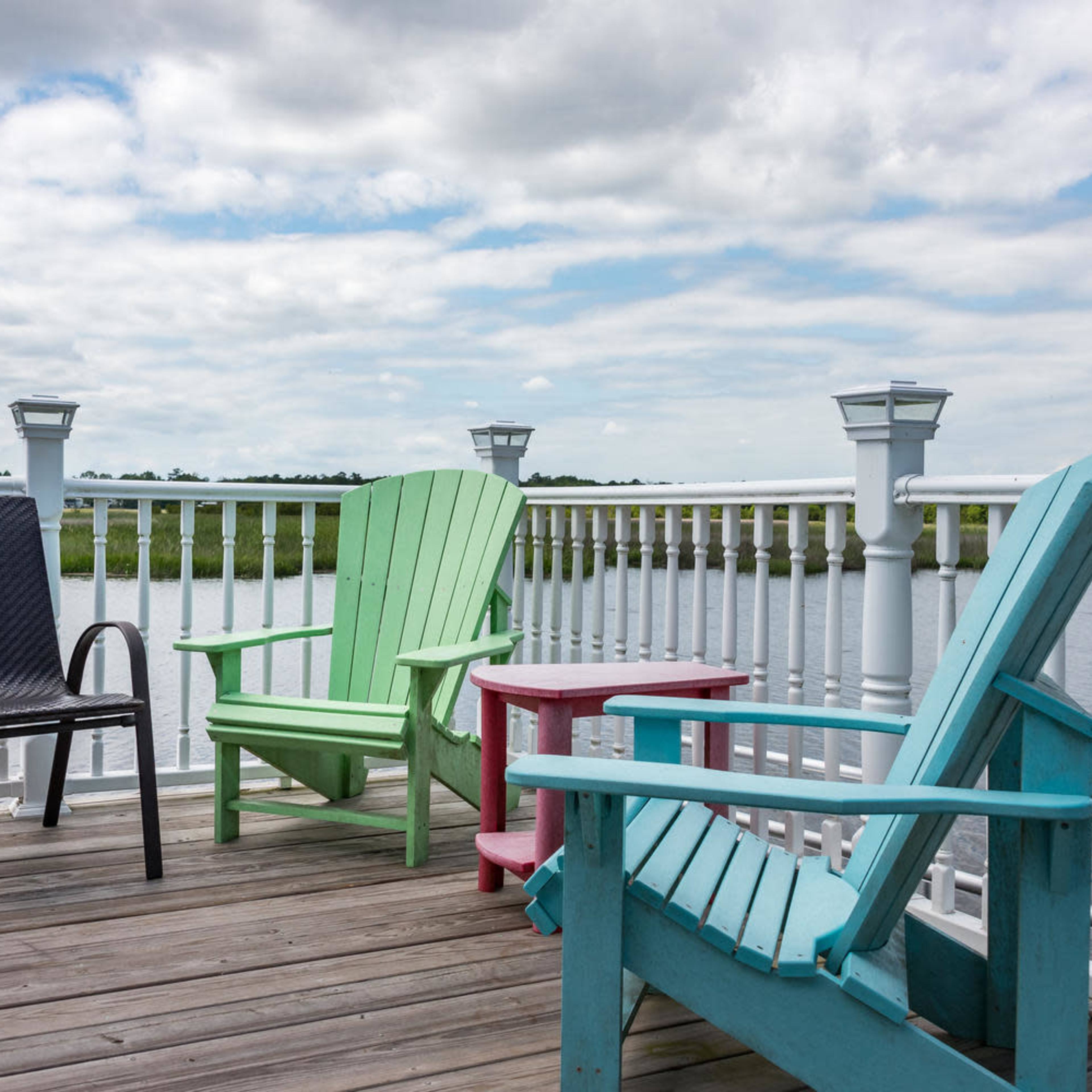 A wooden deck with three colorful chairs facing a calm body of water under a cloudy sky.