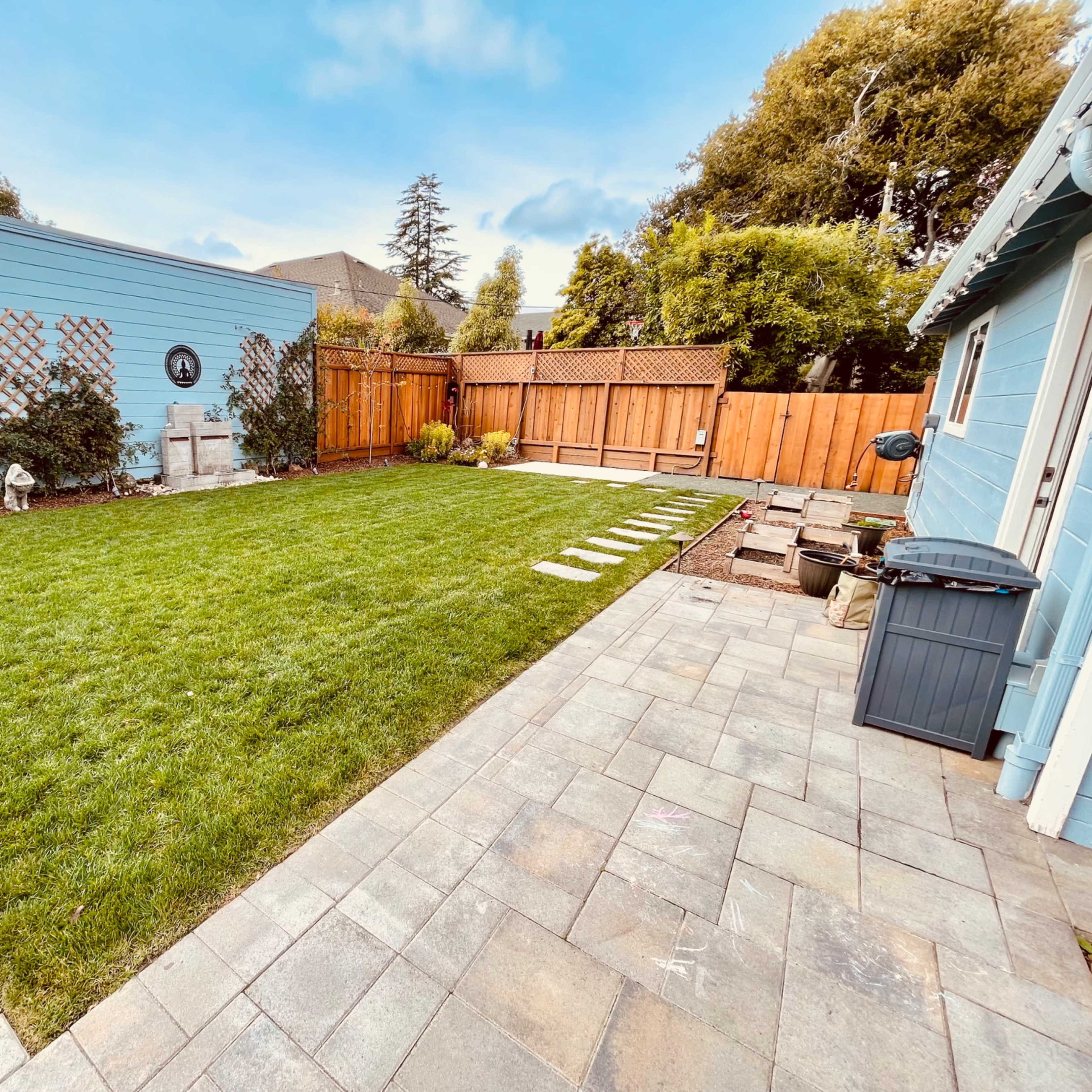 A well-maintained backyard with a green lawn, stone pathway, paved patio areas, and wooden fencing.