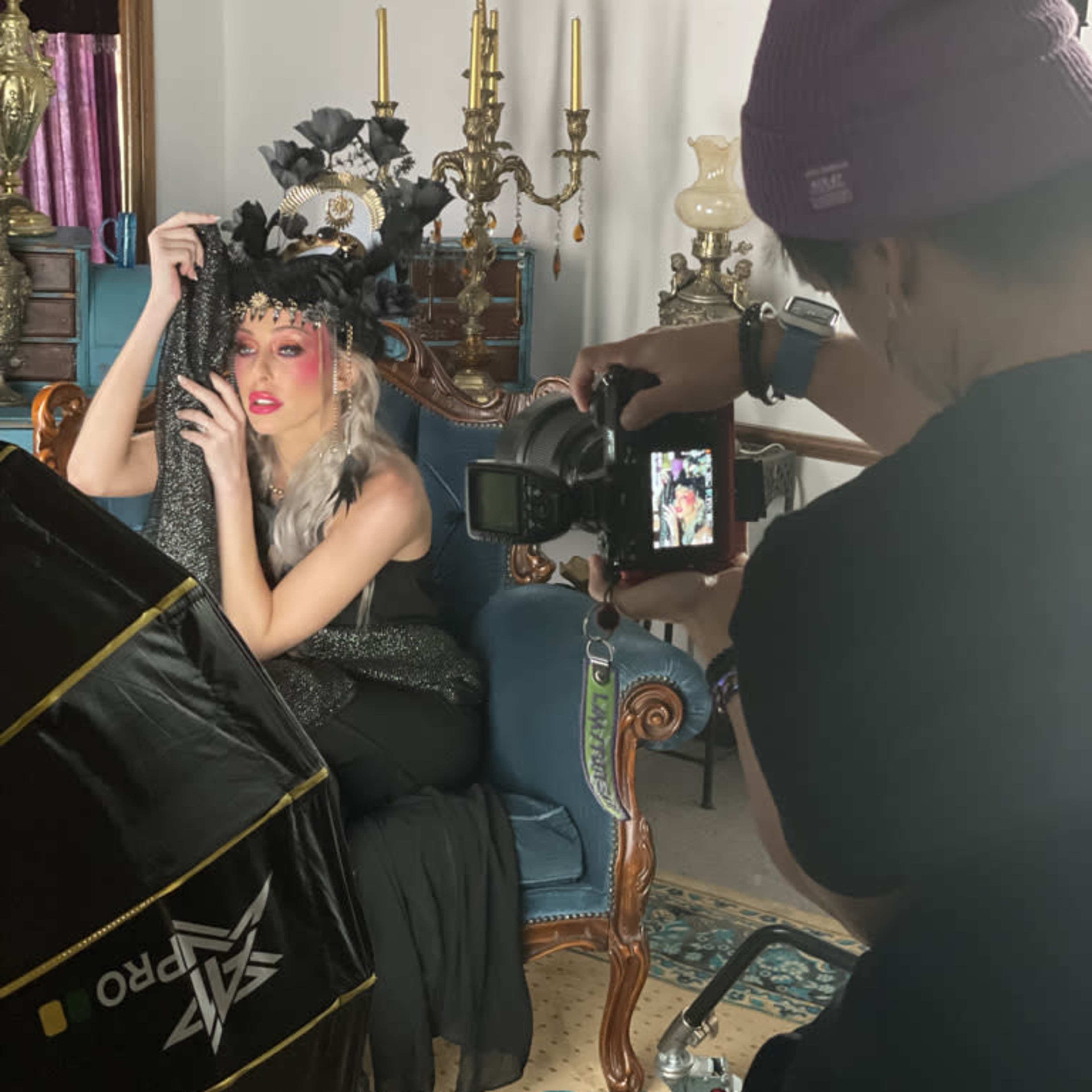 A model poses on a vintage chair while a photographer captures the scene with a camera in a well-decorated interior.