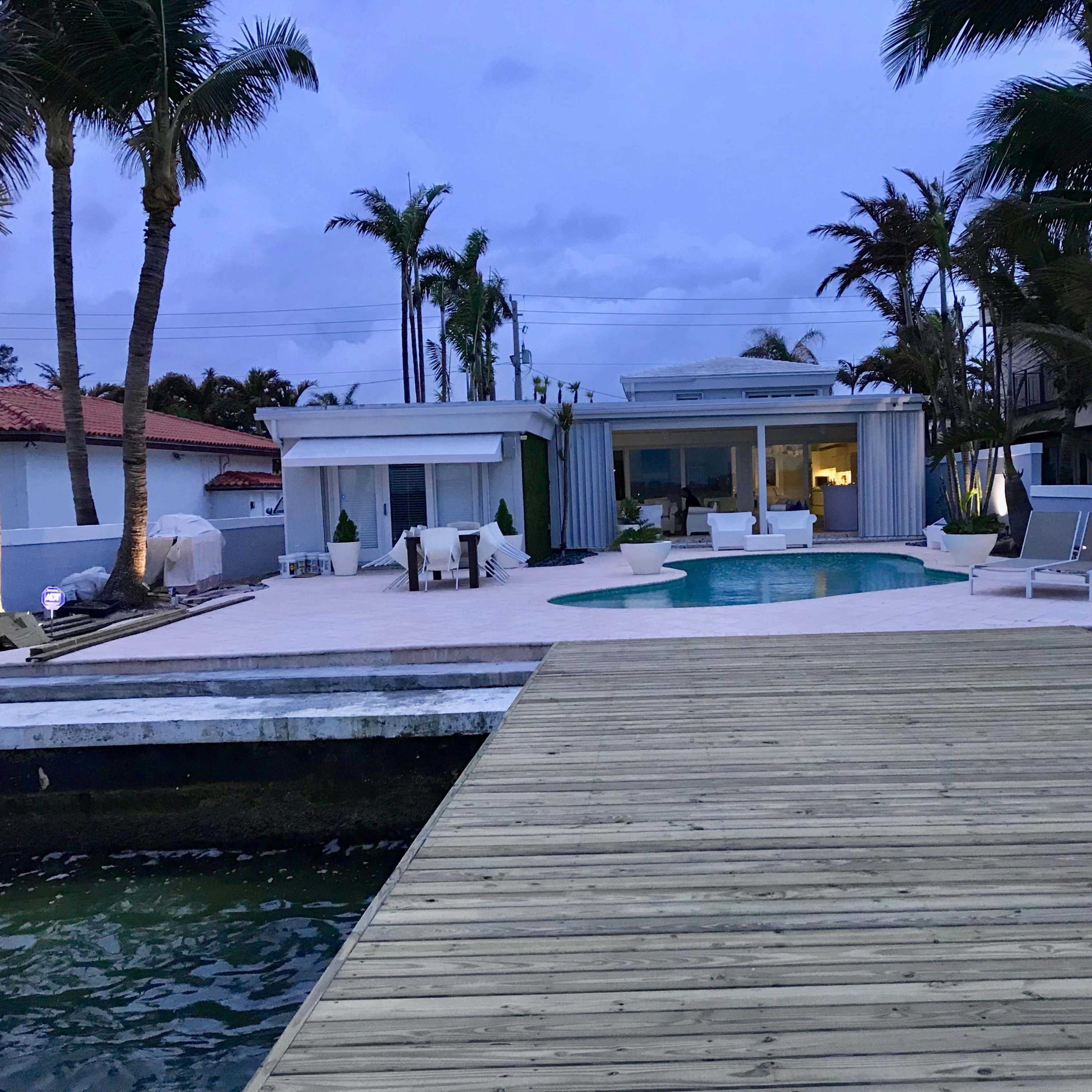 A modern waterfront villa with a pool, surrounded by palm trees and a wooden deck extending over the water.