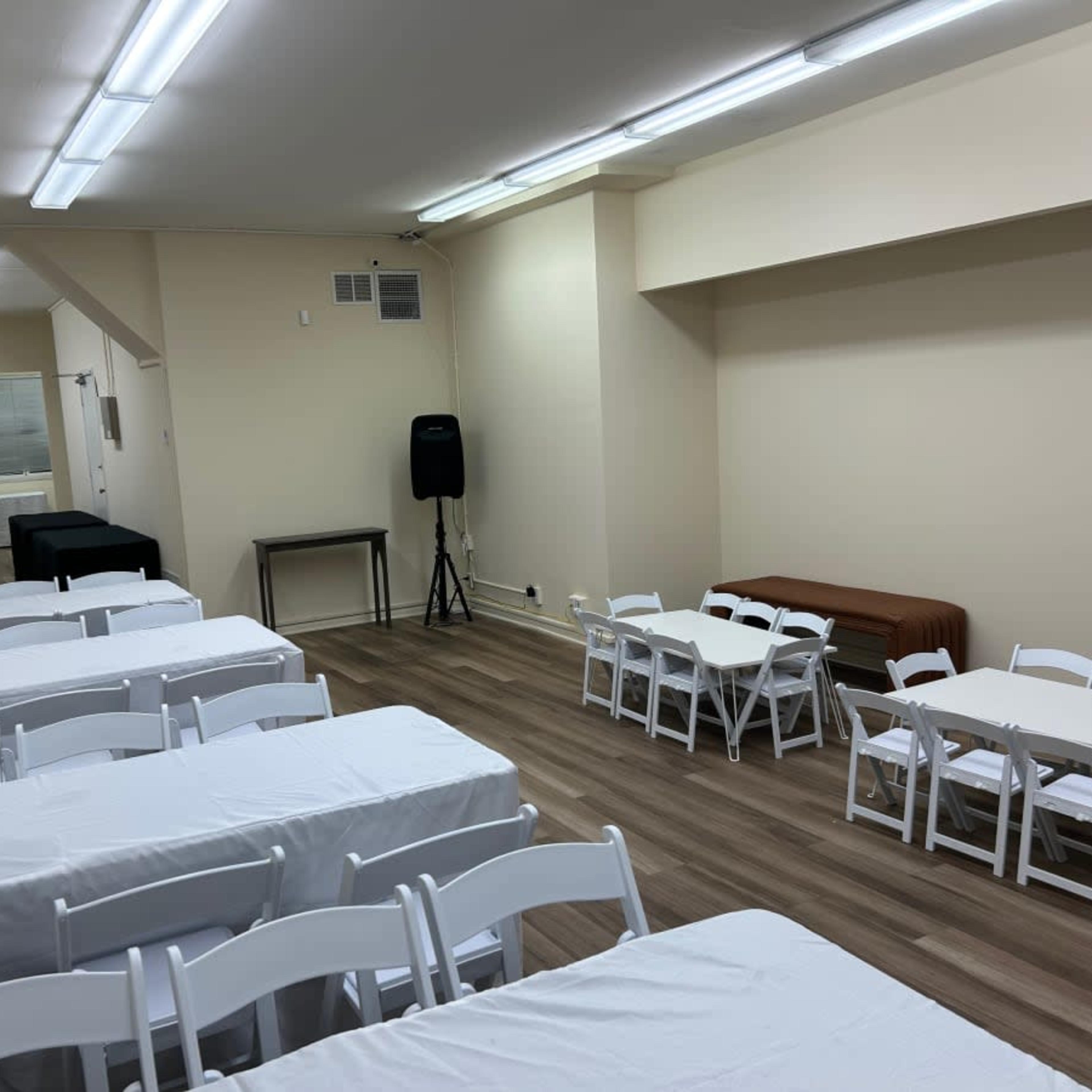 A spacious room with multiple white tables and chairs arranged, including a speaker and a service table along the walls.