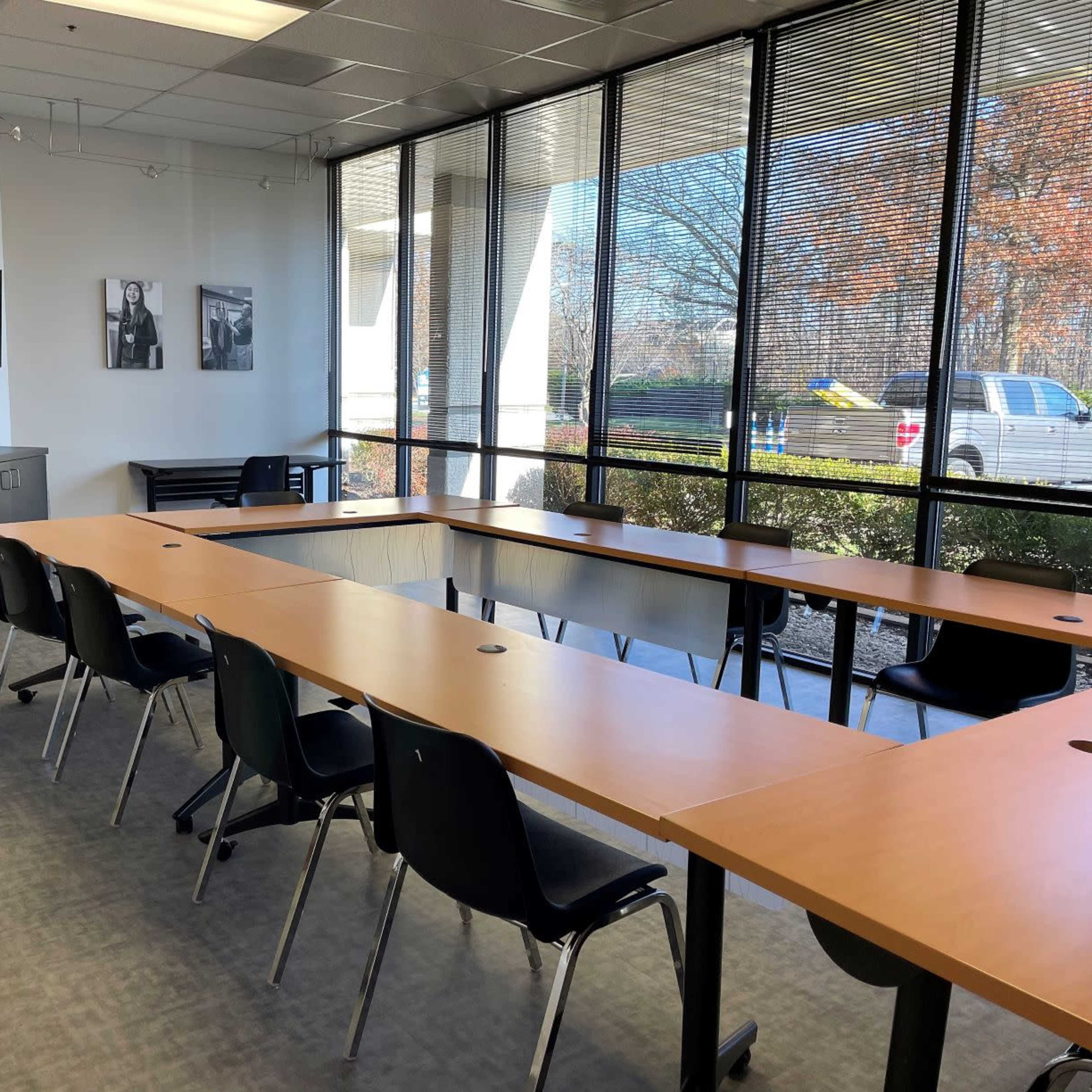 A bright conference room features a long wooden table with black chairs, large windows providing natural light, and a television mounted on the wall.