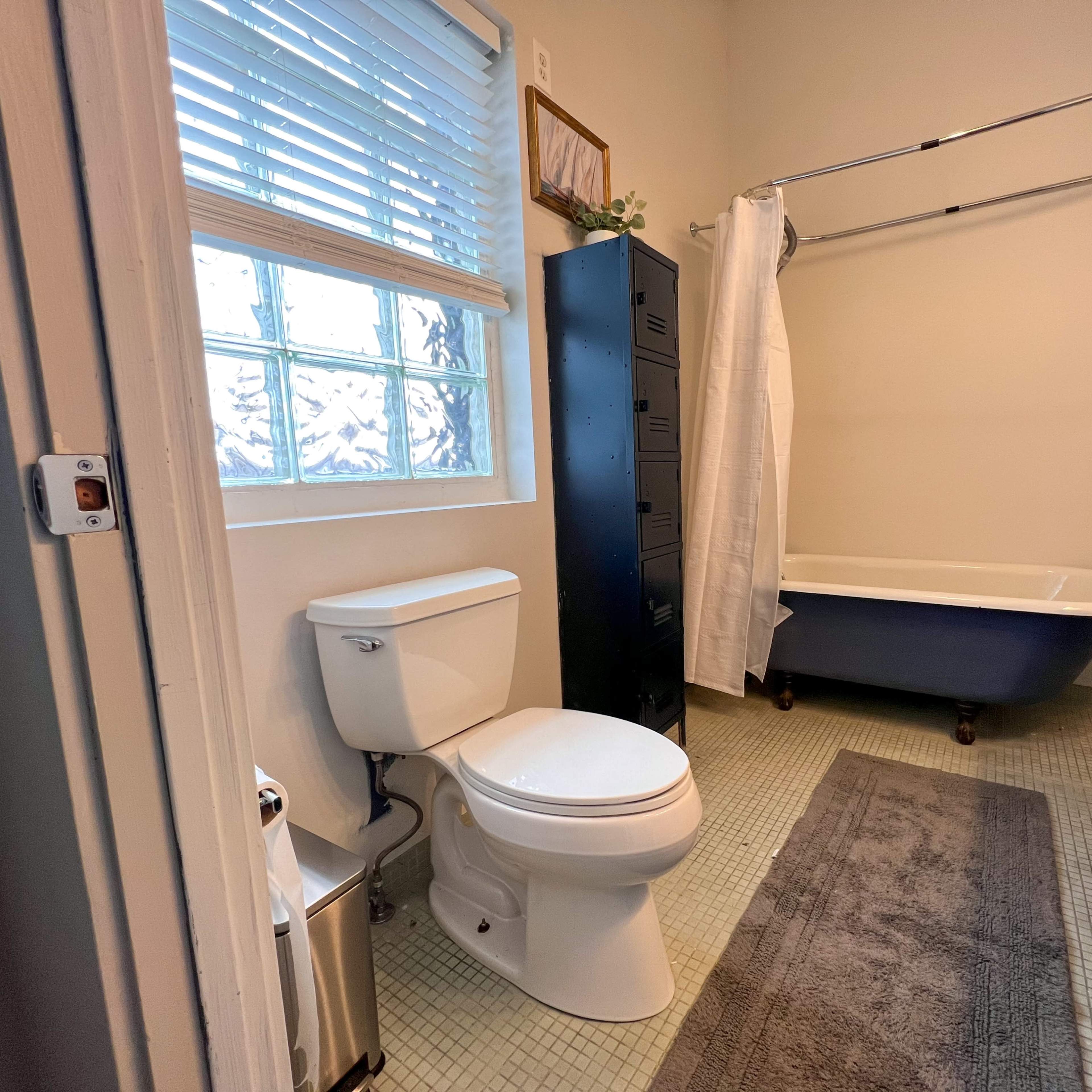 A bathroom features a clawfoot tub, a toilet, a sink with dark cabinetry, and a window with blinds.