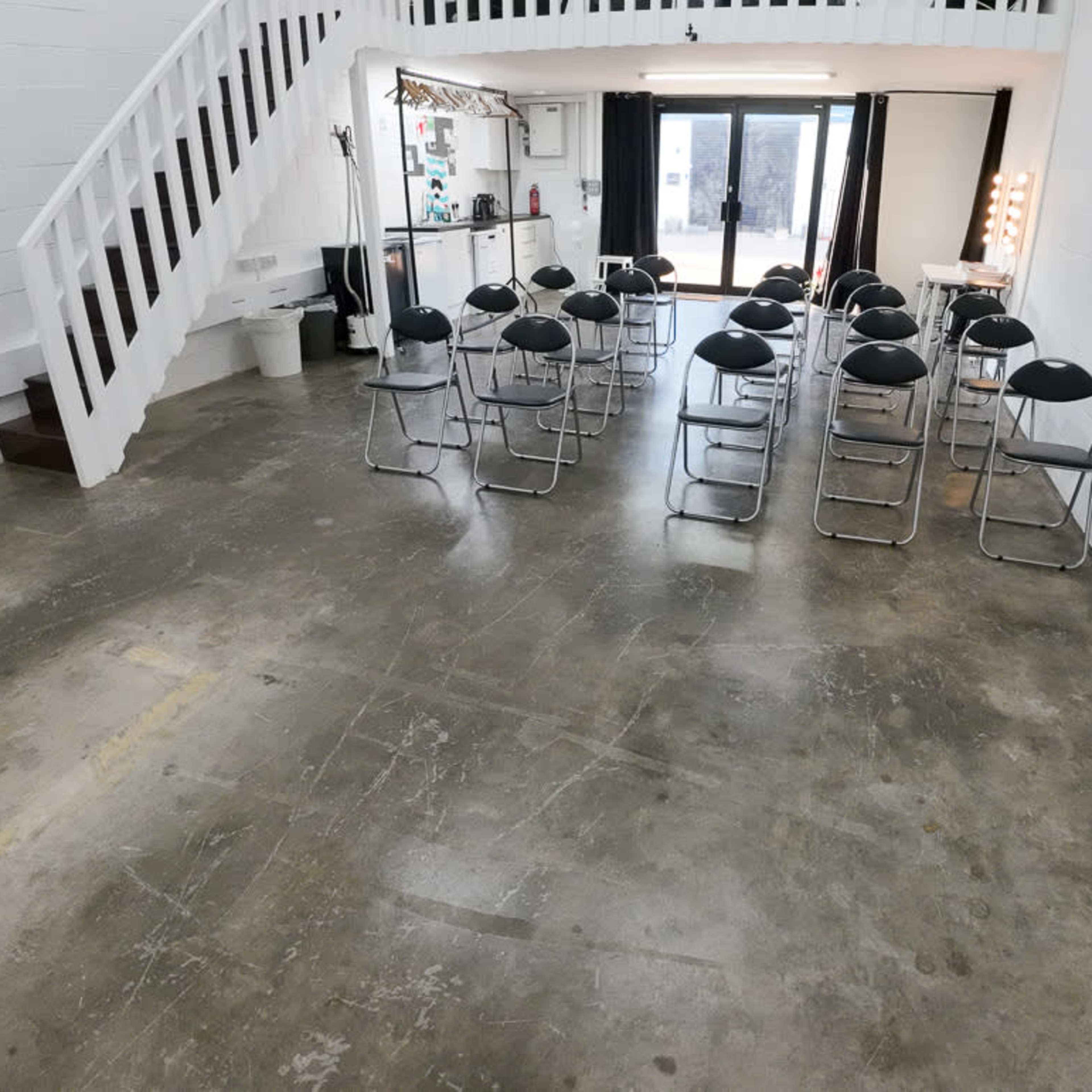 A large, empty room with a polished concrete floor and a row of black folding chairs arranged in the center, adjacent to a staircase and large windows.