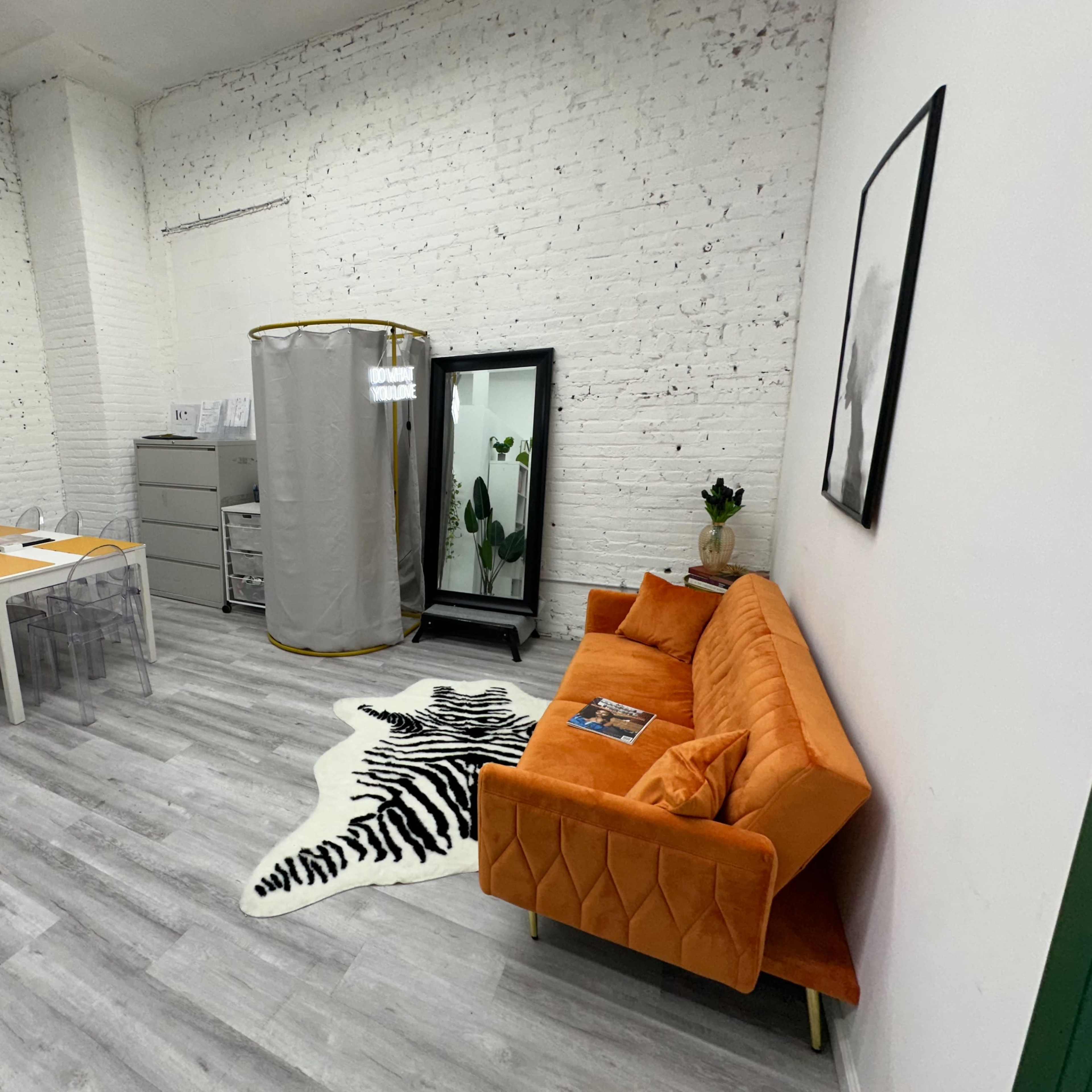 A modern office space features an orange sofa, a glass dining table, a clothing rack, and potted plants against a backdrop of white brick walls.