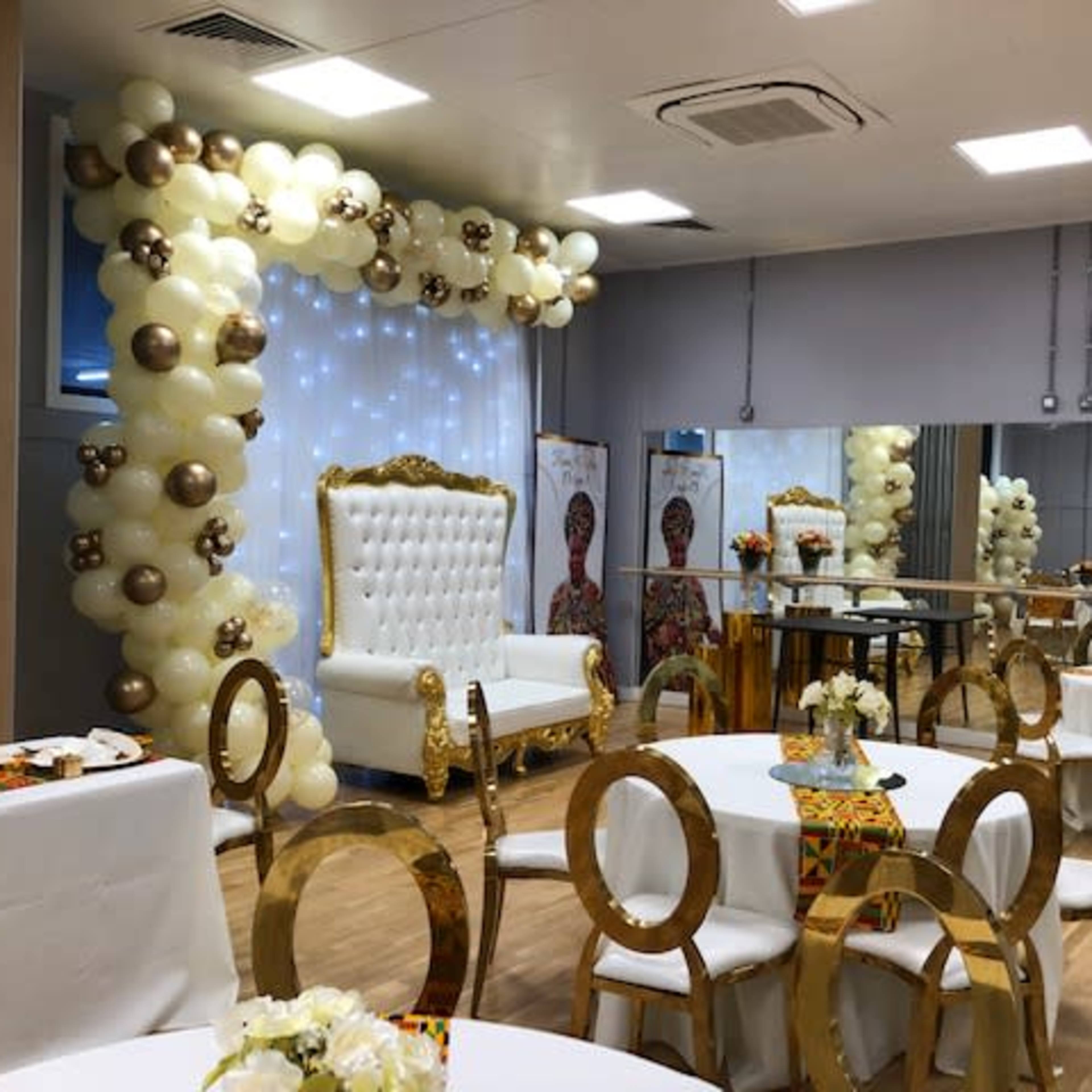 A decorated event space with a white and gold themed seating area, featuring a large white tufted chair and a backdrop of balloons.