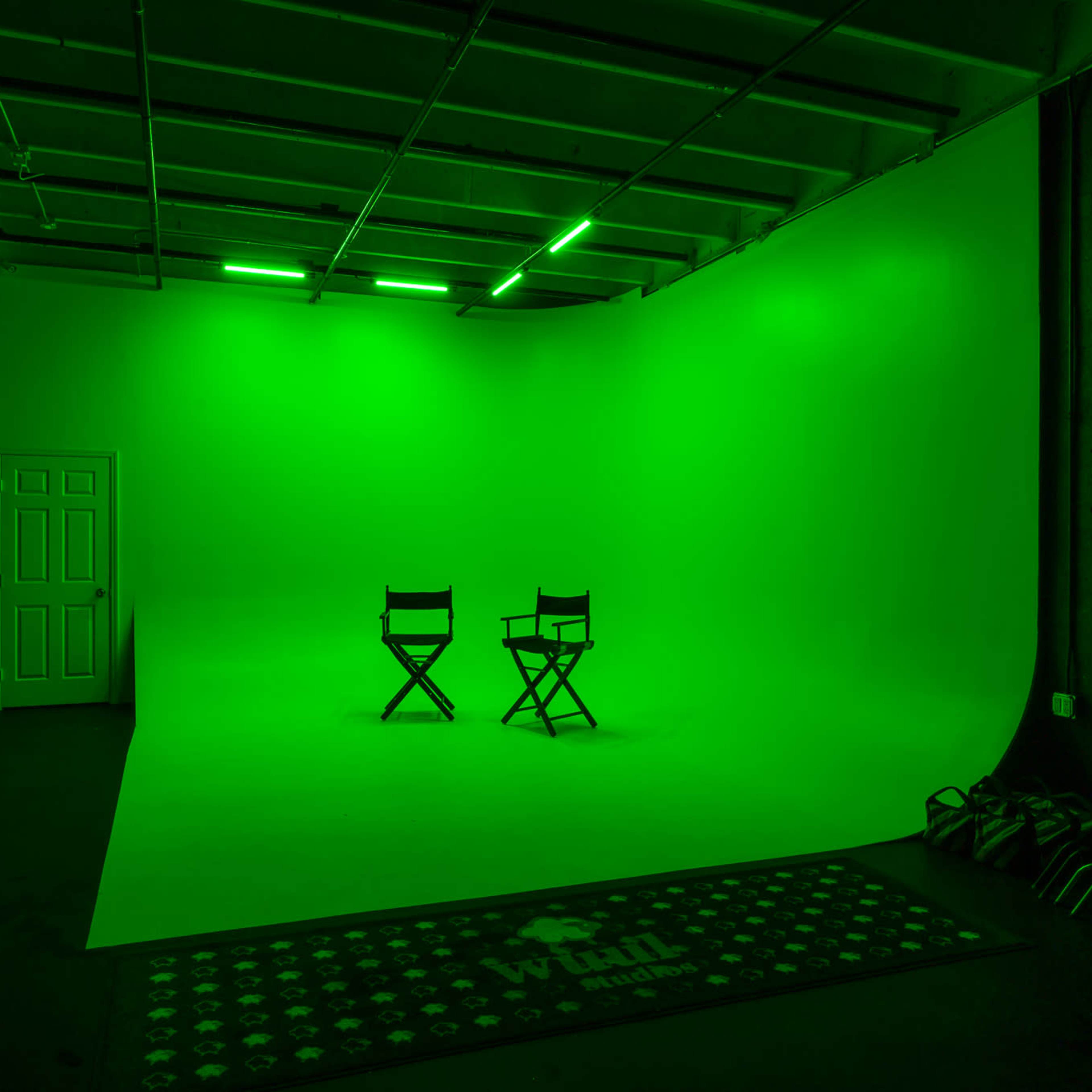 An empty studio space with two director's chairs positioned in front of a green backdrop and illuminated by green lights.