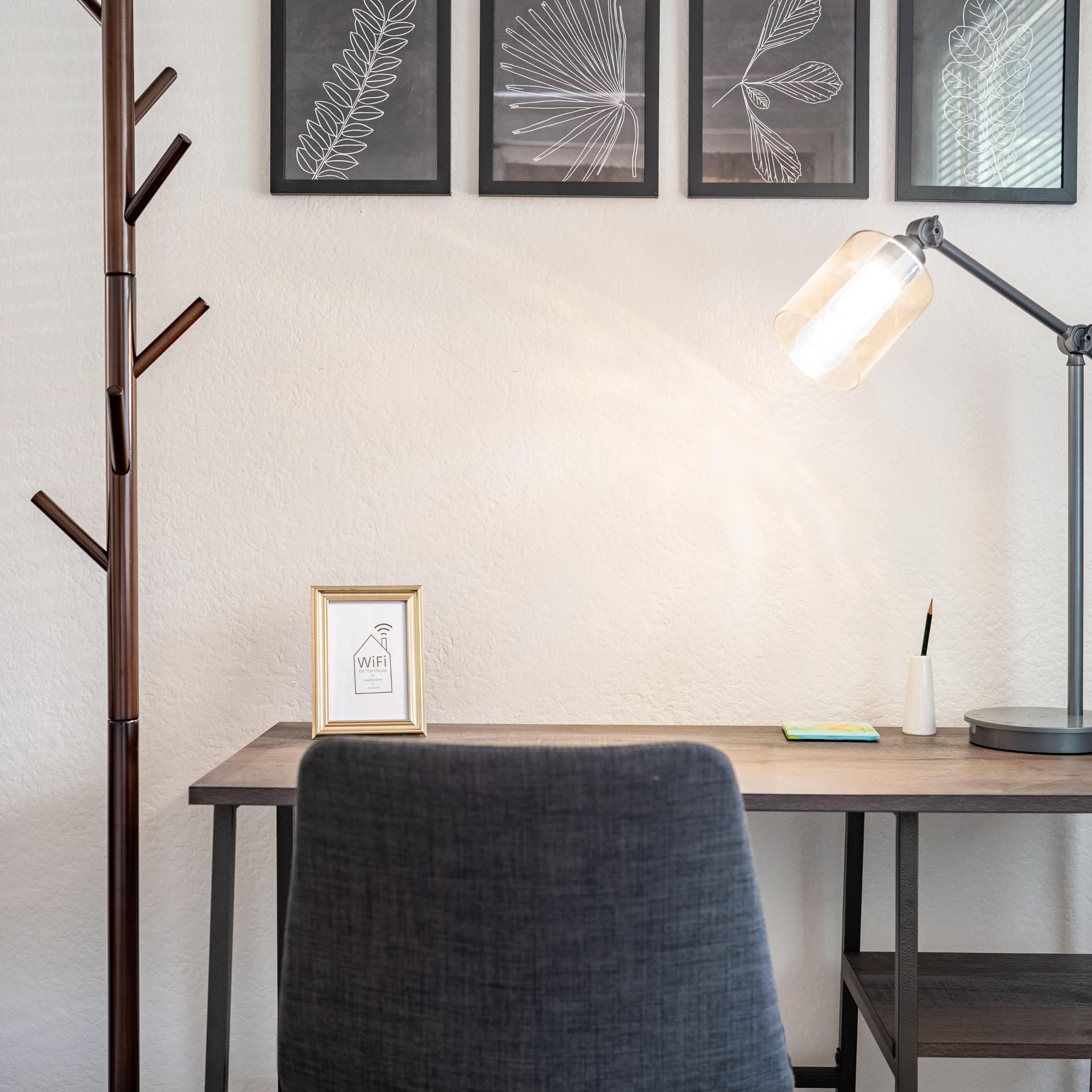 A minimalist workspace features a wooden desk with a lamp, a decorative picture frame, and a chair, accompanied by a coat rack and wall art above.