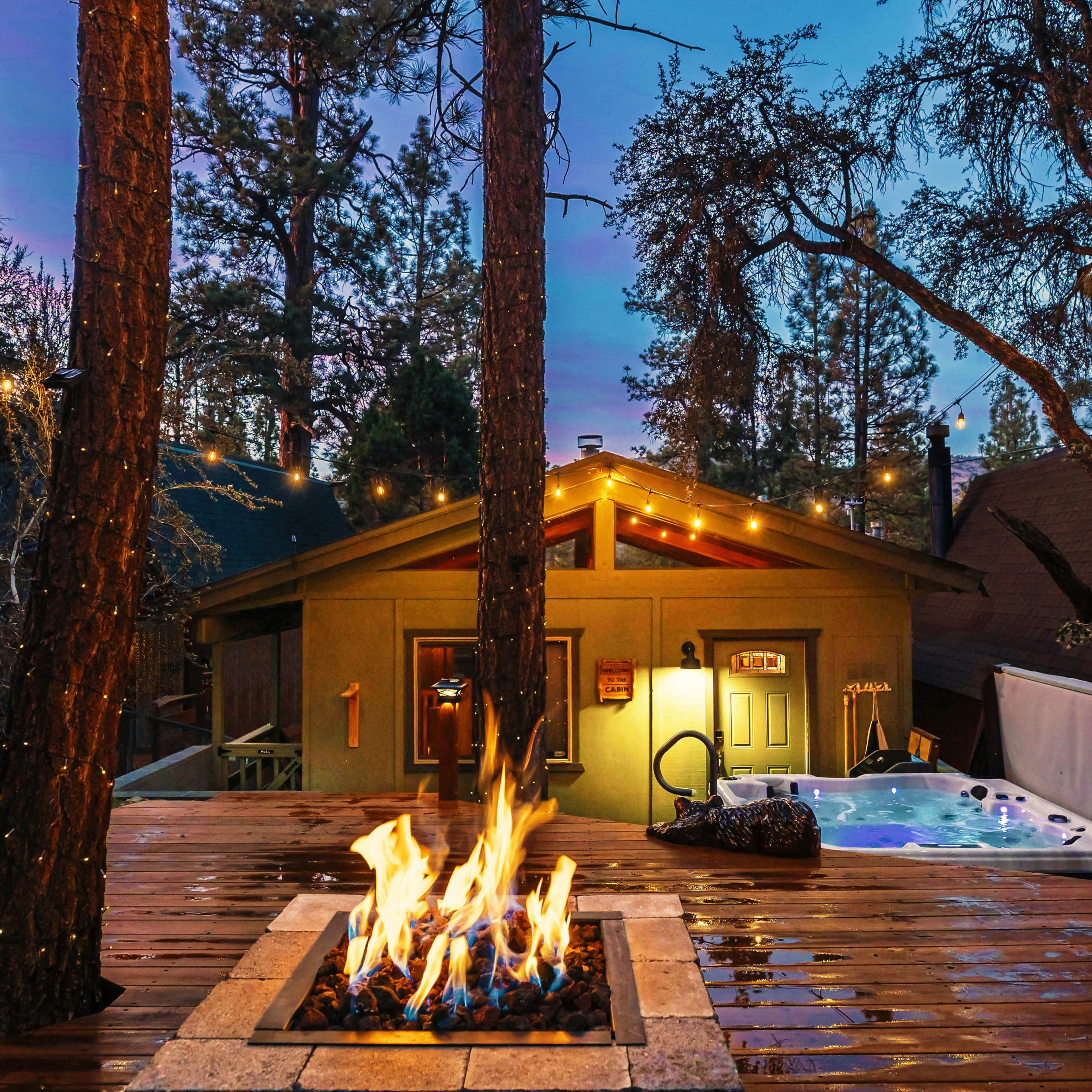 A fire pit surrounded by wooden decking is illuminated by string lights and nearby trees, with a hot tub visible in the background.