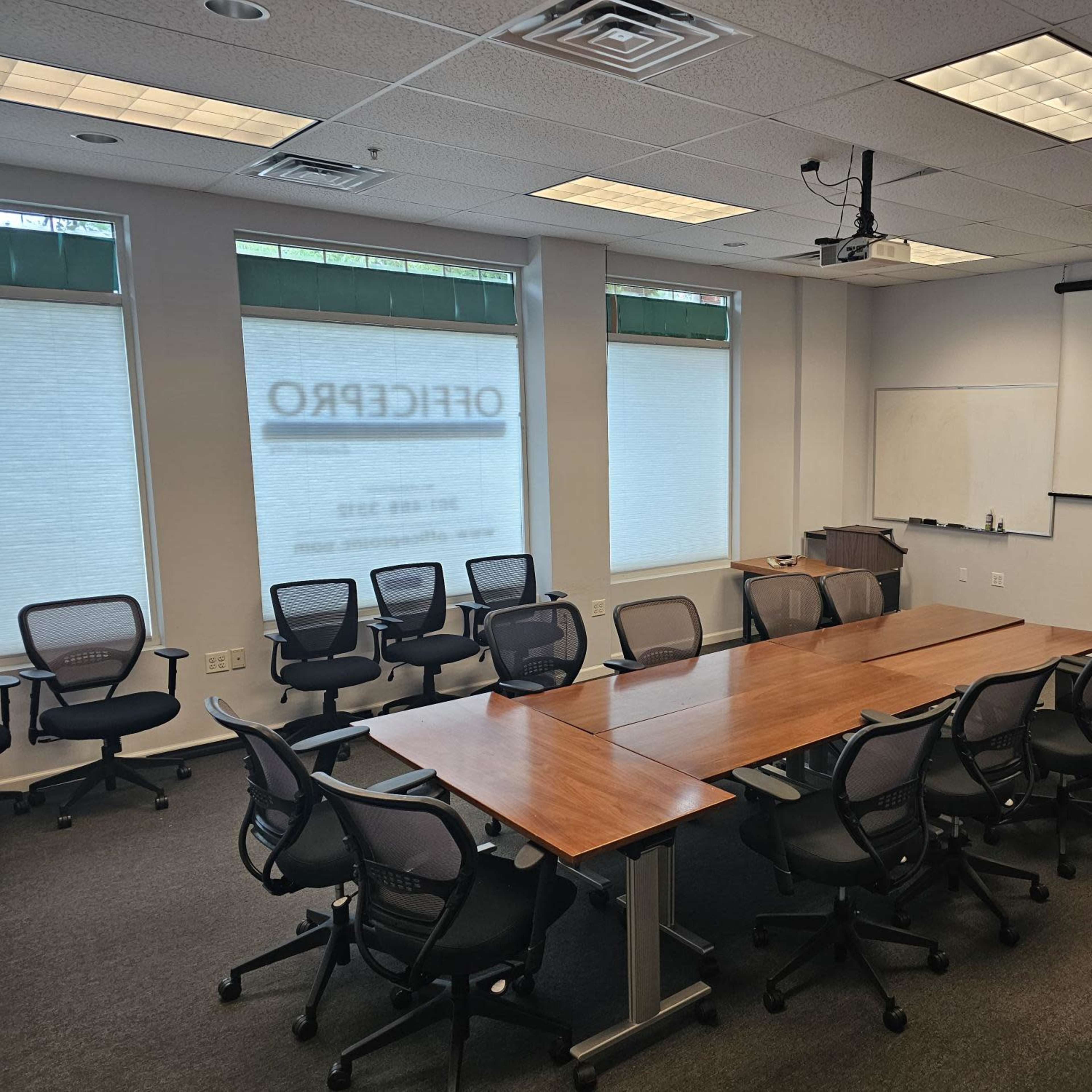 A conference room with a long wooden table surrounded by chairs, a projector mounted on the ceiling, and windows with blinds.