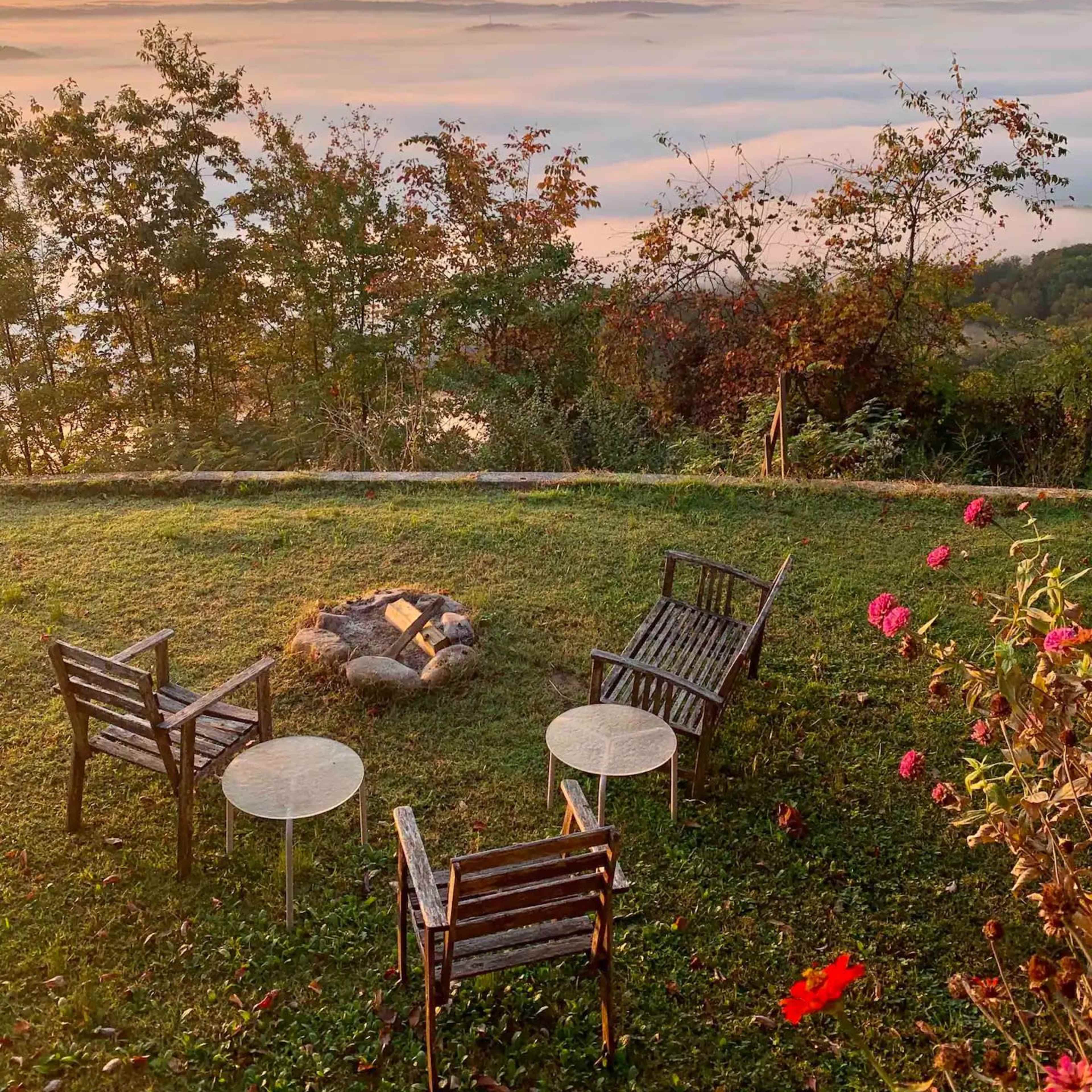 A set of four wooden chairs and two small tables is arranged around a stone fire pit on a grassy area overlooking a misty landscape at sunrise.