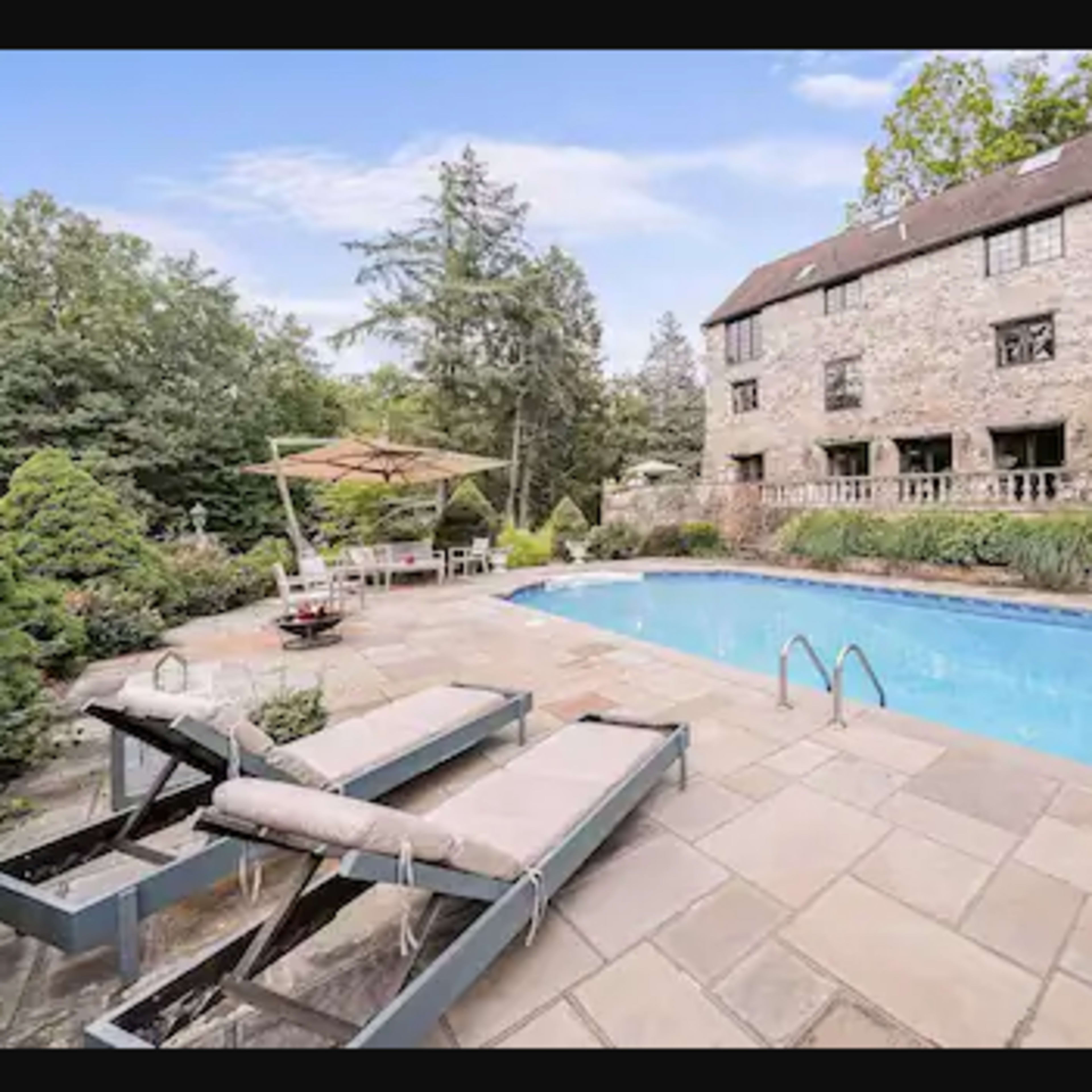 A stone house with a swimming pool surrounded by a stone patio, lounge chairs, and lush greenery.