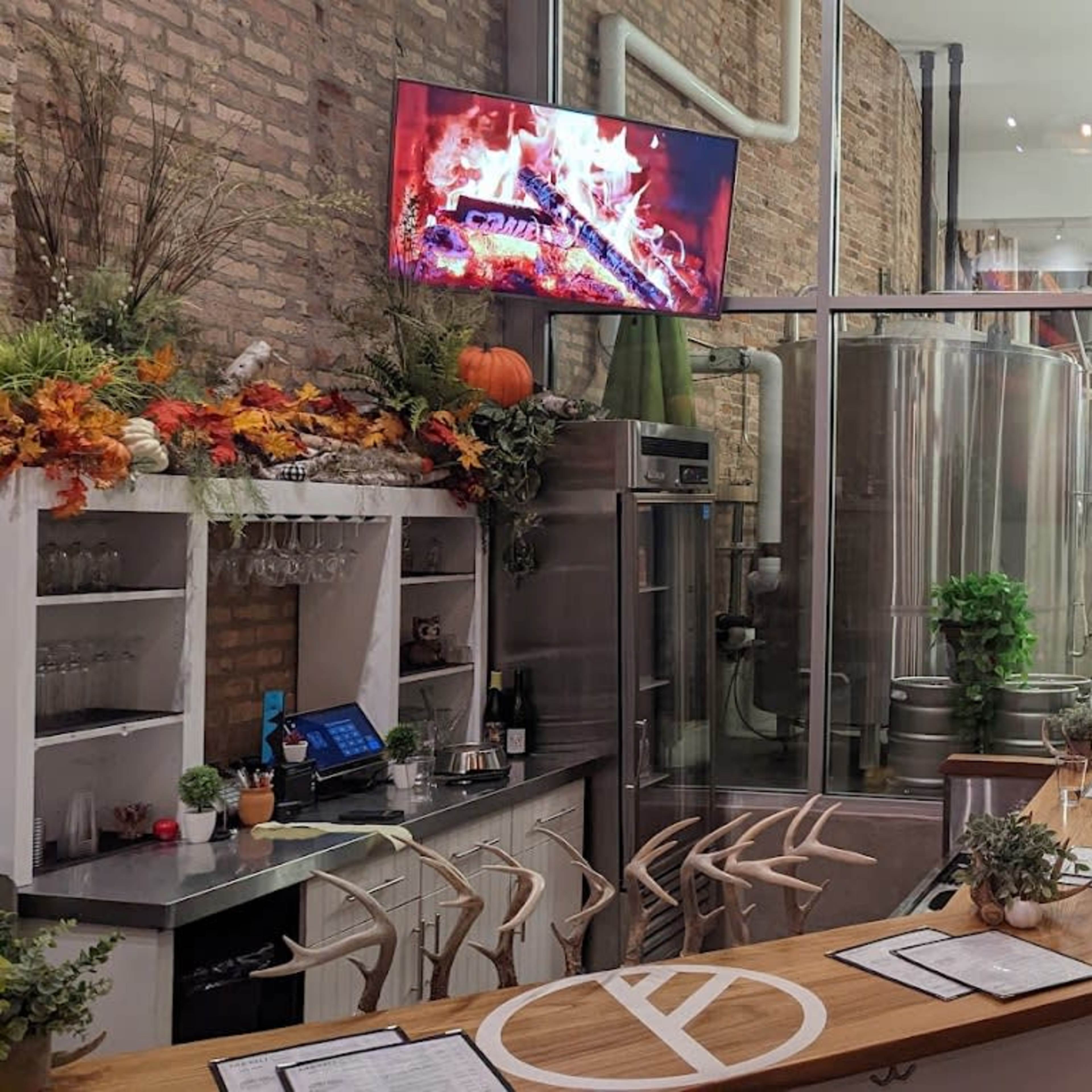 A modern brewery interior with a wooden bar counter, stainless steel brewing equipment in the background, and a large screen displaying a fire simulation.