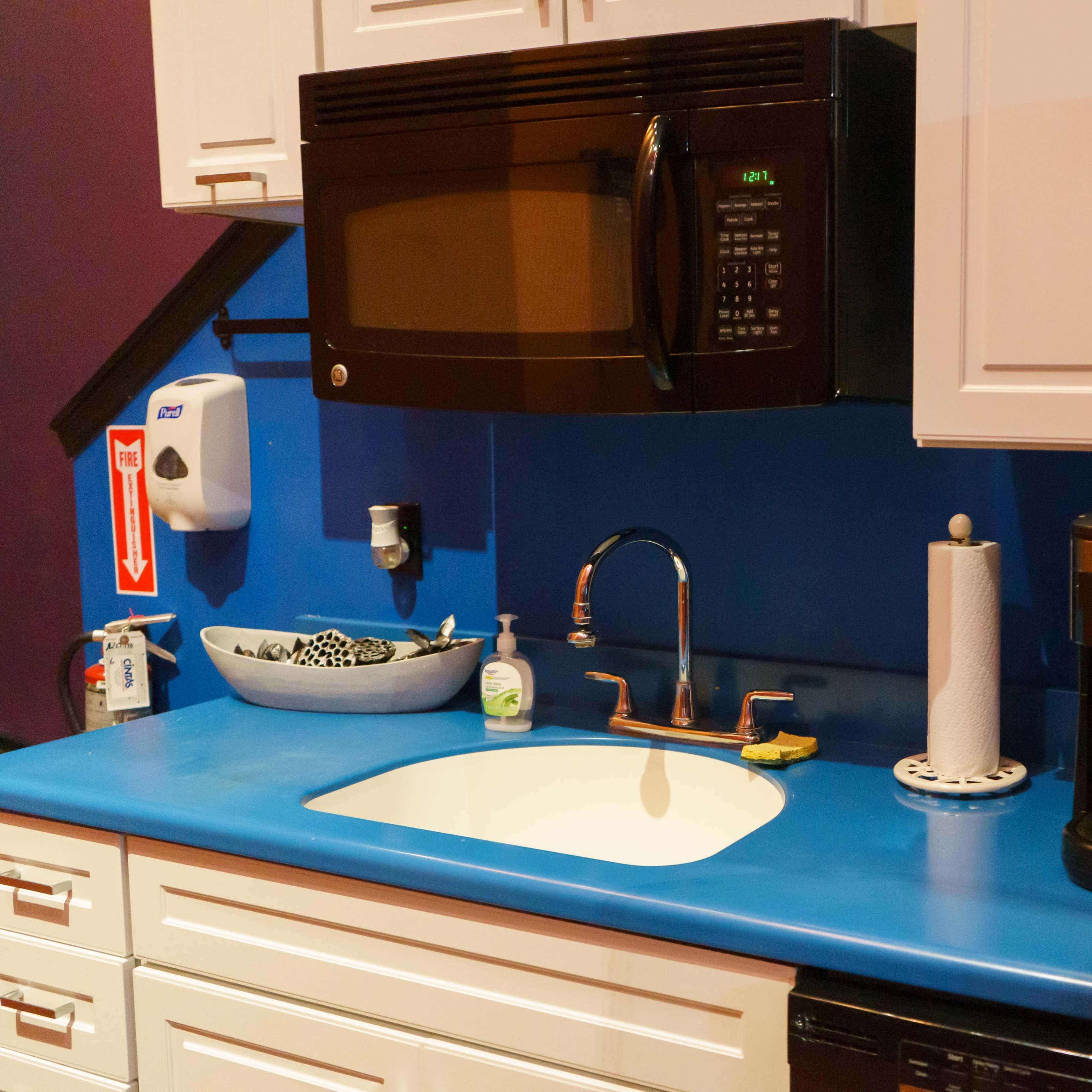 A kitchen countertop featuring a microwave, sink, coffee maker, and various kitchen items against a blue wall.