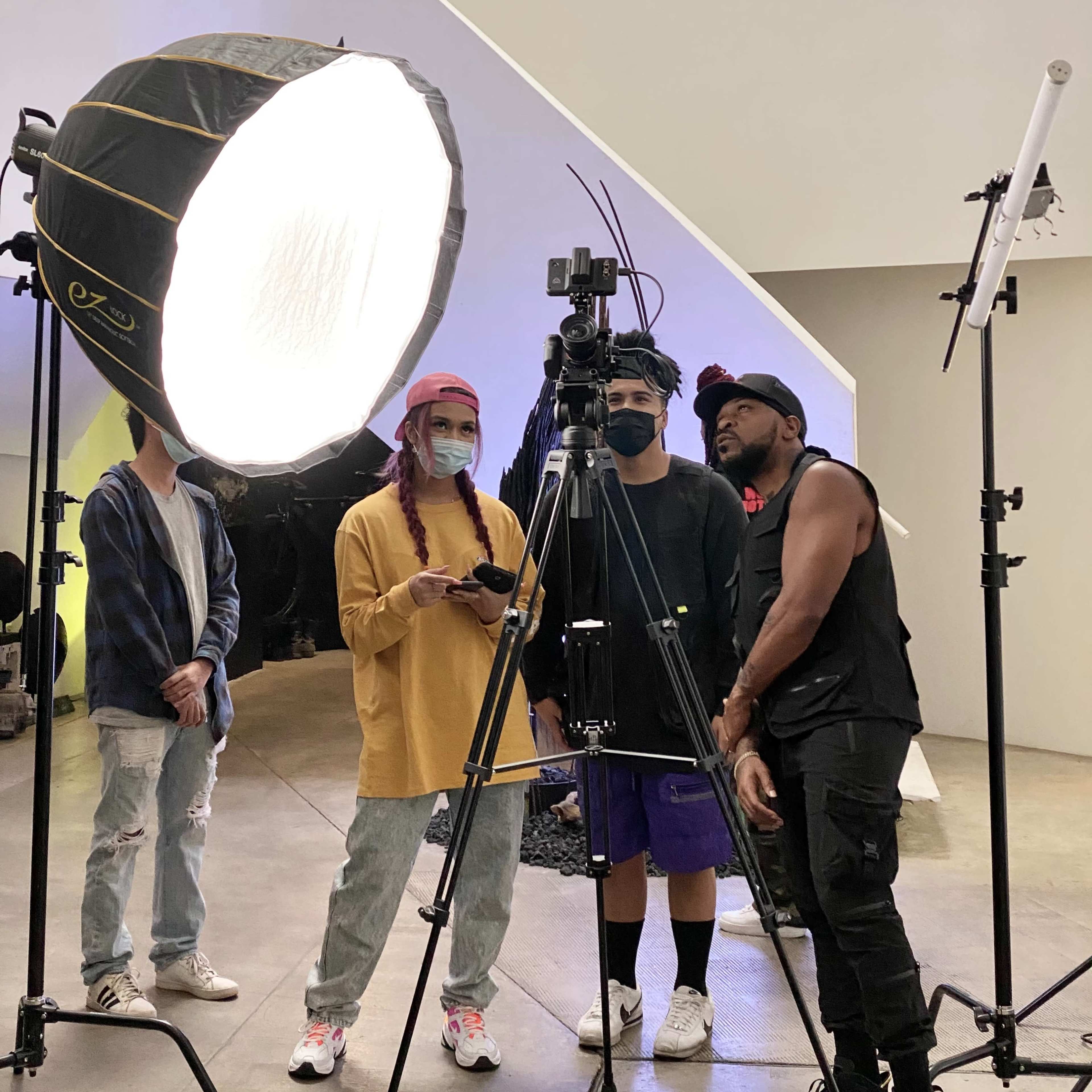 A group of four individuals is gathered around a camera setup, with one person adjusting the equipment while others observe in a spacious indoor studio.