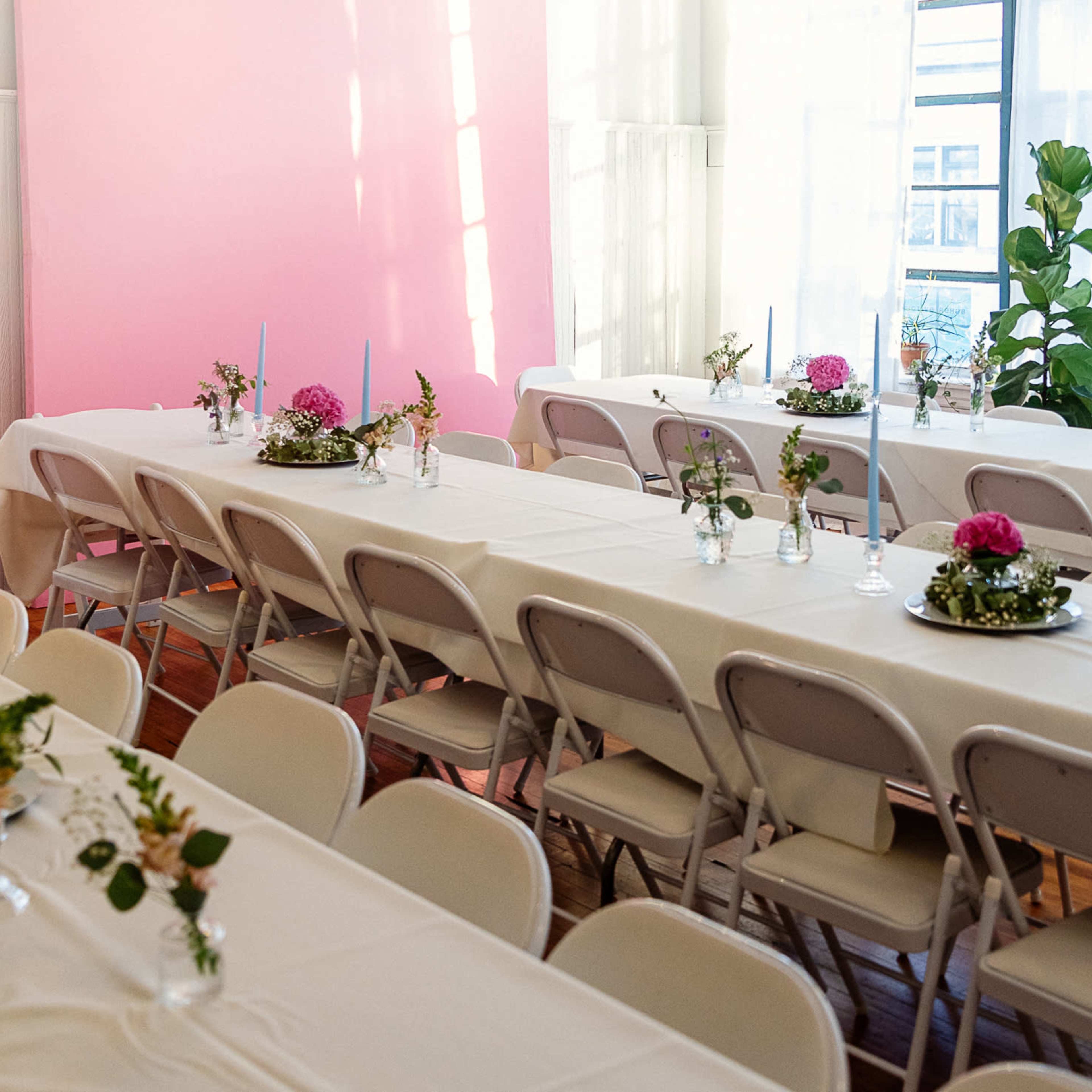 A spacious dining area features long tables covered with white tablecloths, arranged with floral centerpieces and candles, alongside a pink wall and large windows.