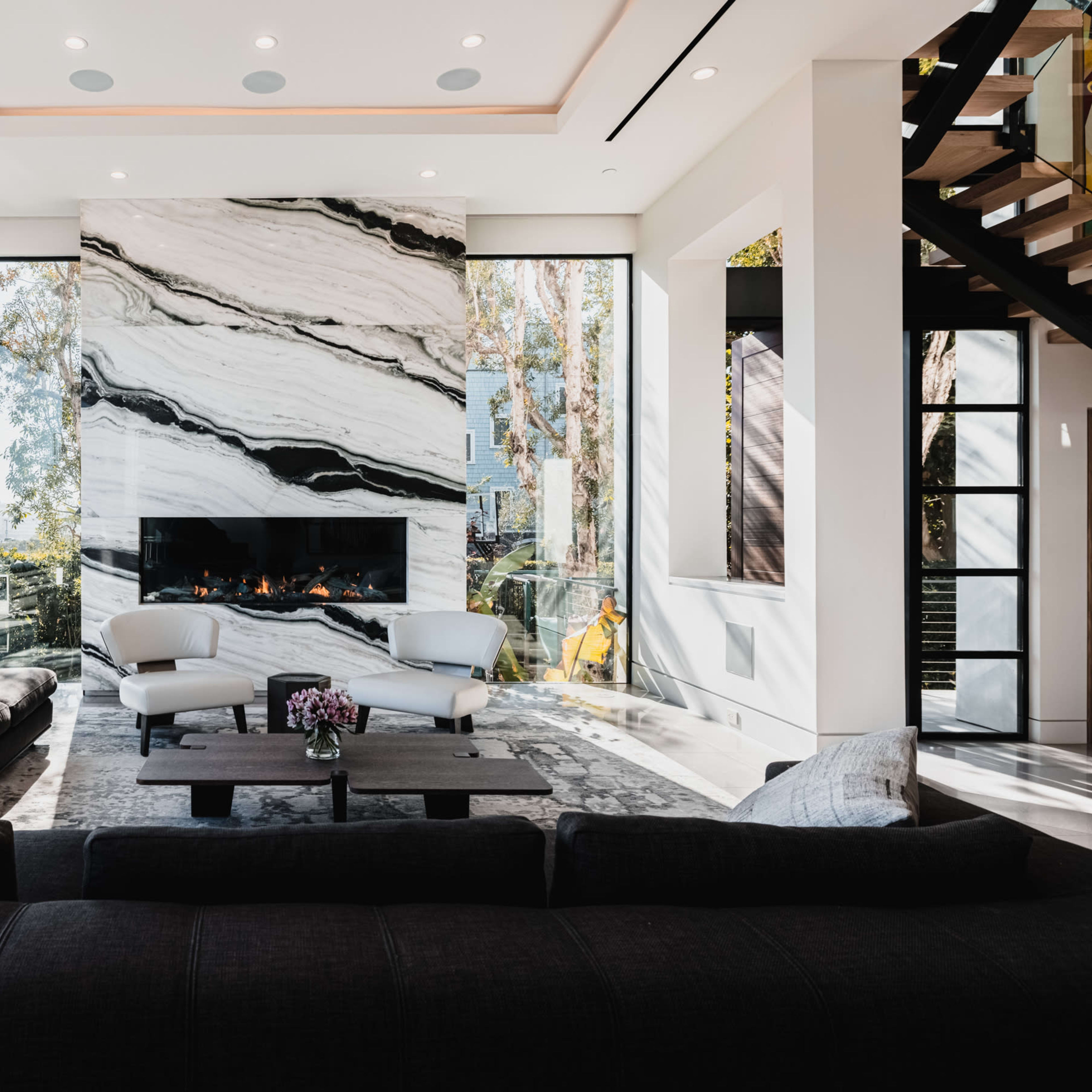 A modern living room features a large marble fireplace, floor-to-ceiling windows, and a sectional sofa arranged around a coffee table.