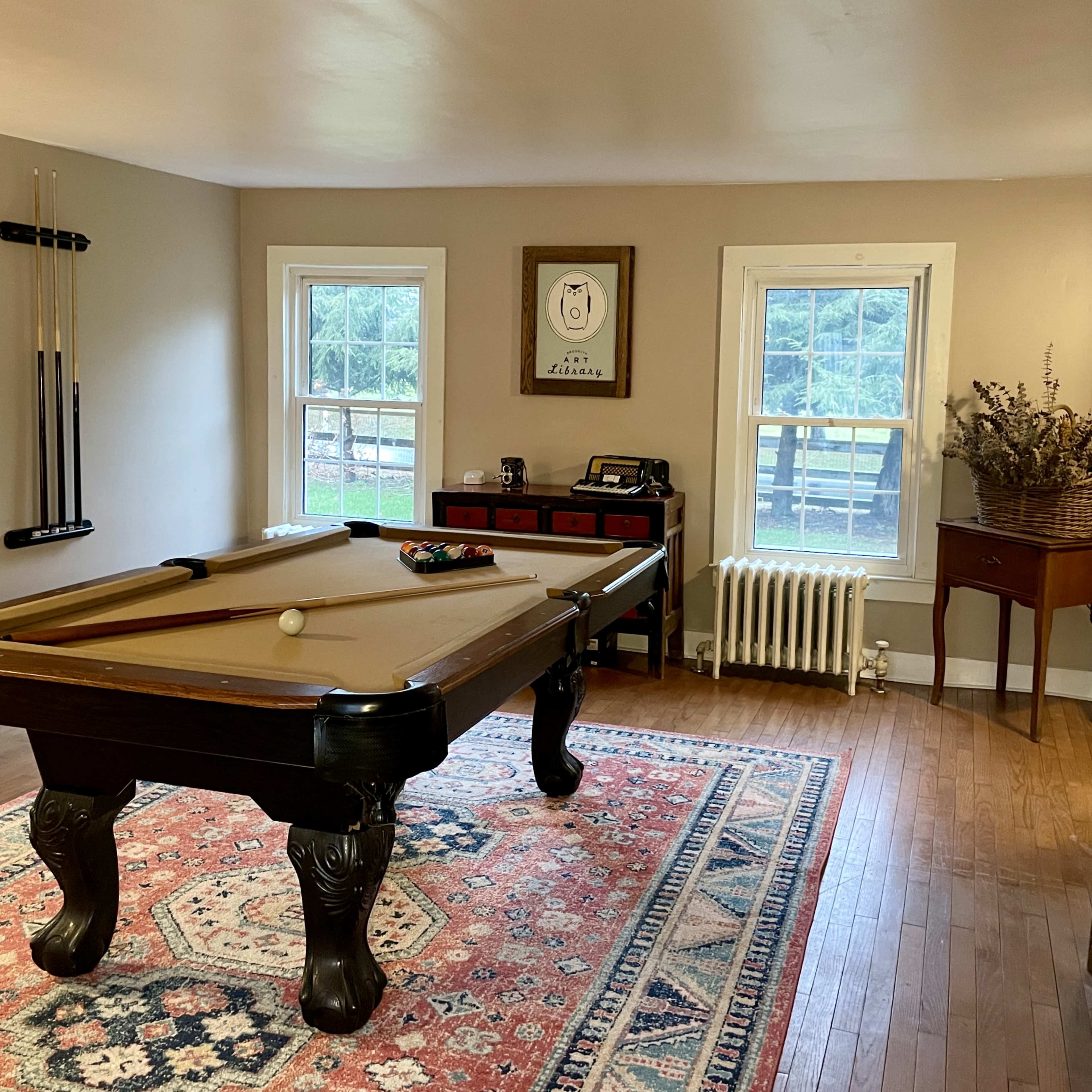 A room with a pool table, two windows allowing natural light, a small side table, and a vintage-style cabinet against the wall.