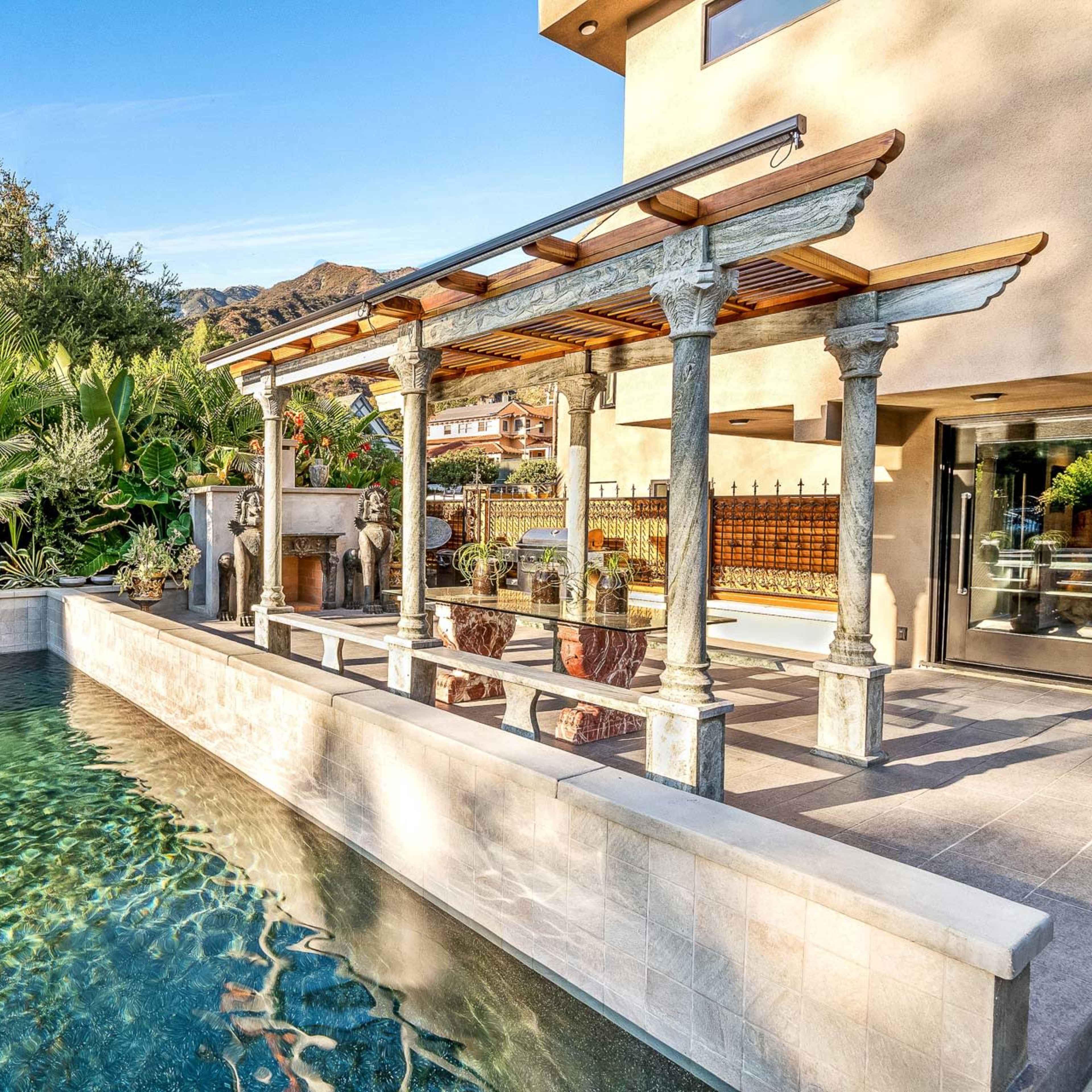 A clear swimming pool with a stone edge is surrounded by lush greenery and features a wooden pergola with columns.