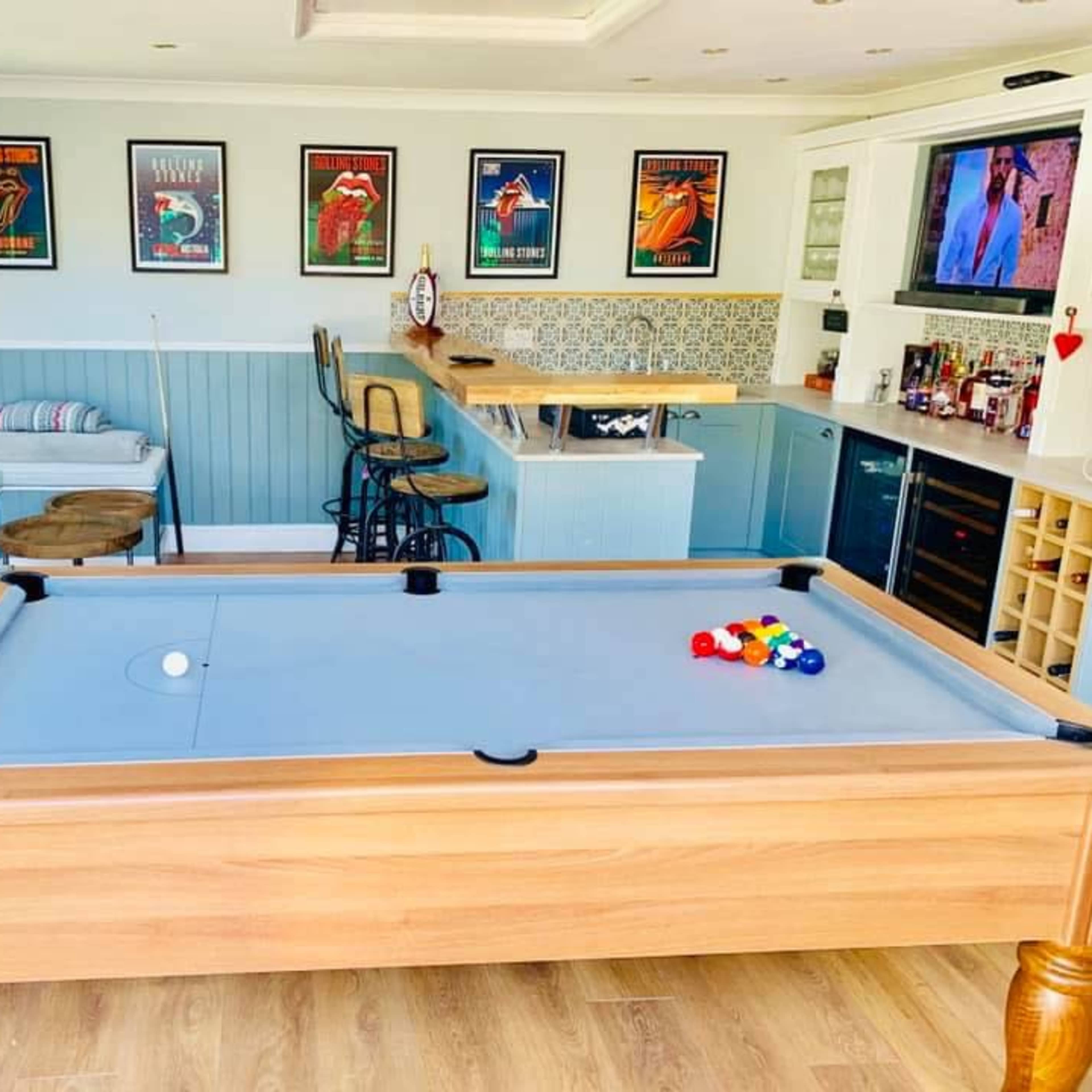 A brightly lit game room featuring a pool table in the center, surrounded by a bar area, a seating nook, and decorated walls with framed posters.