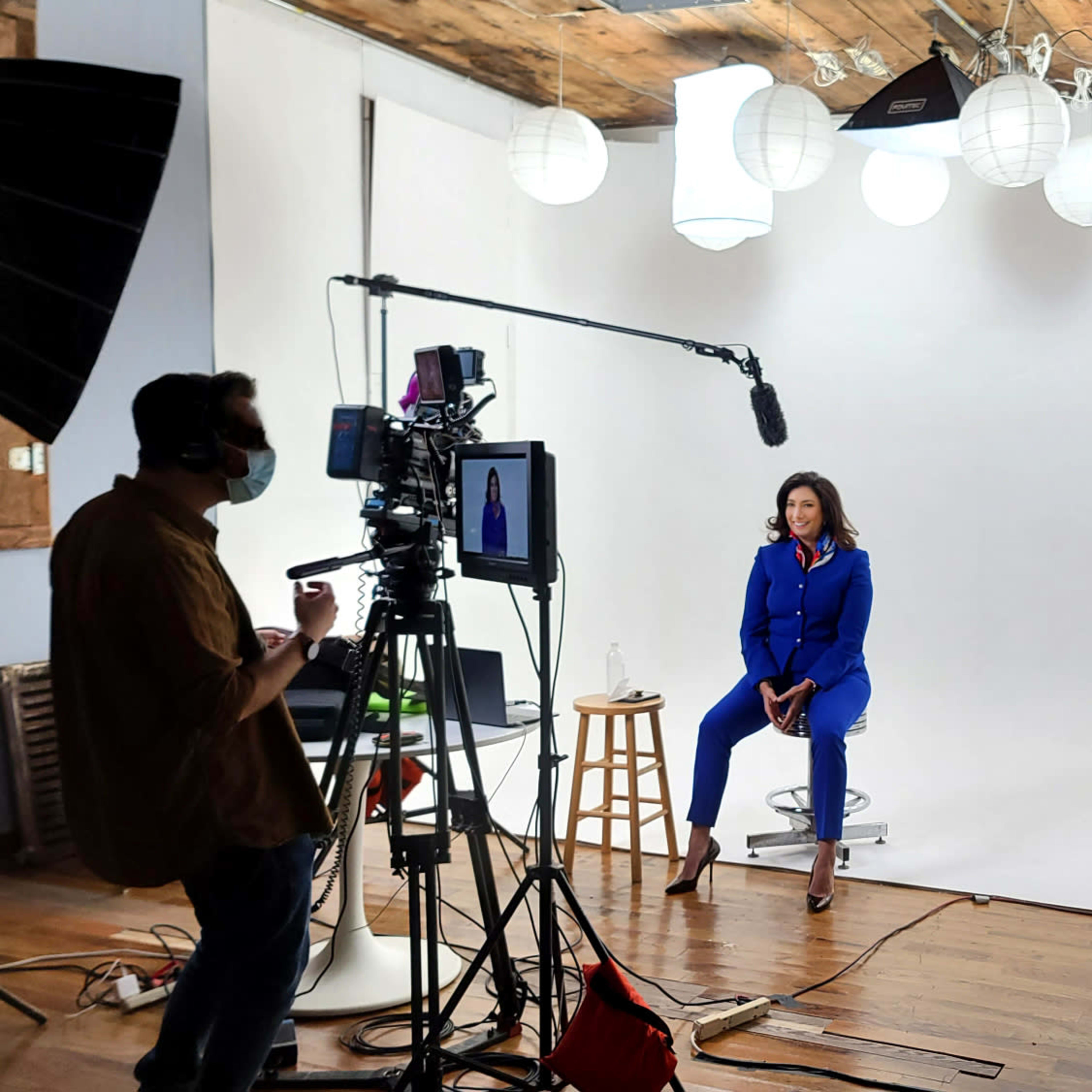 A woman in a blue suit sits on a stool in front of a camera setup in a brightly lit studio with softbox lights and paper lanterns overhead.