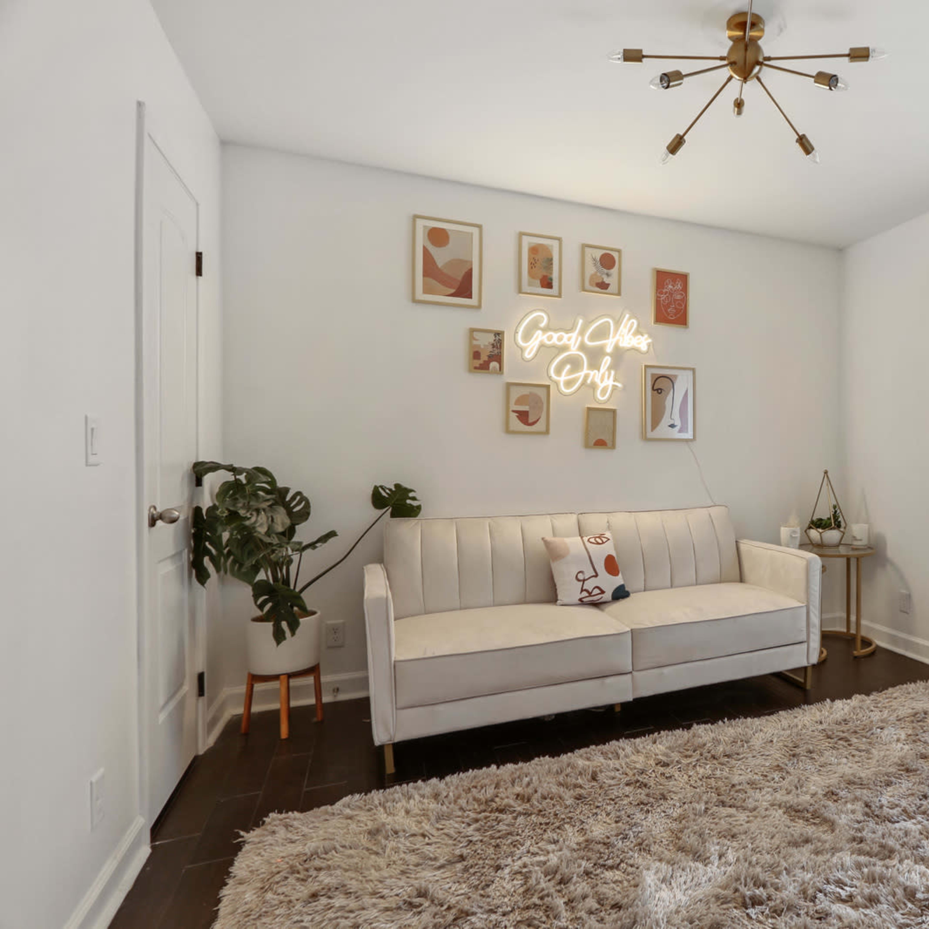 The room features a white sofa, a decorative wall with framed art and a neon sign, and a large area rug on a dark wooden floor.